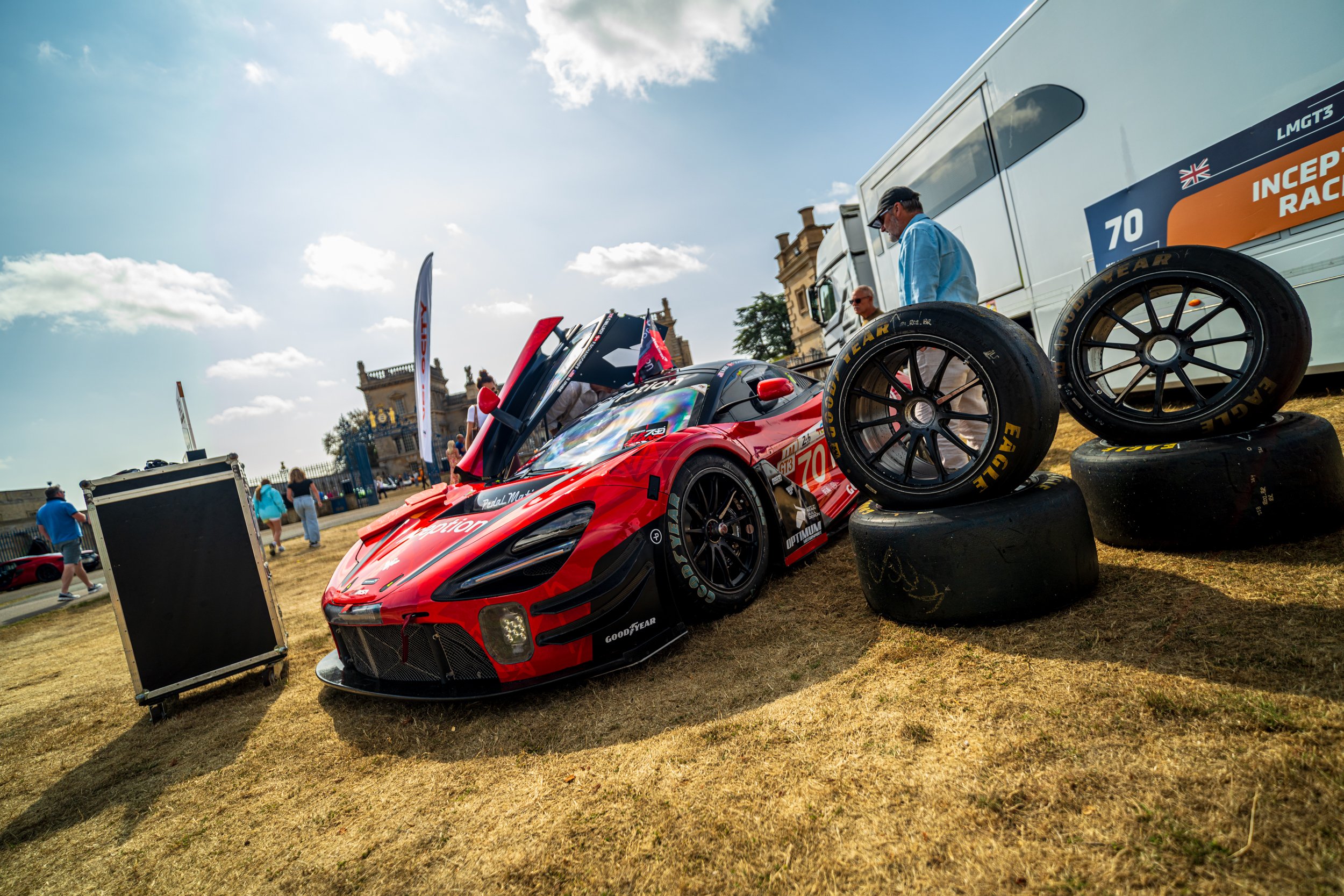 A racing car with a red and black design, number 70, is on display outdoors during a racing event. It is surrounded by spare tires, and spectators are walking around. There are tents and flags in the background, with a historic building and partly cl