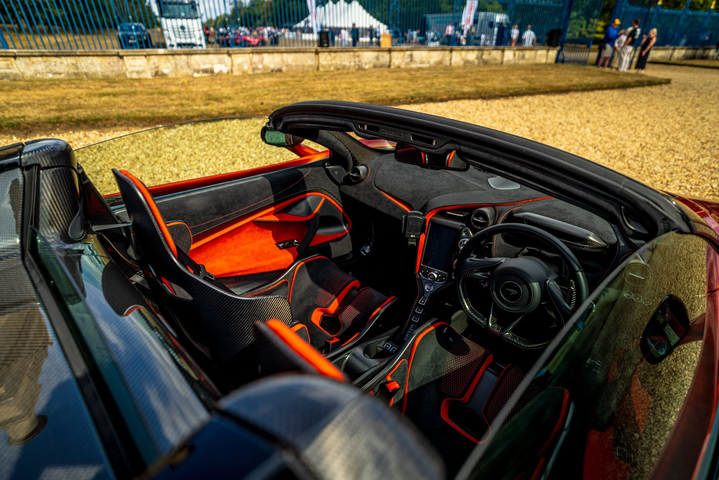 Interior of a sleek, high-performance sports car with black and orange accents, showing the dashboard, steering wheel, and racing seats, set against a backdrop of outdoor event with people and vehicles.