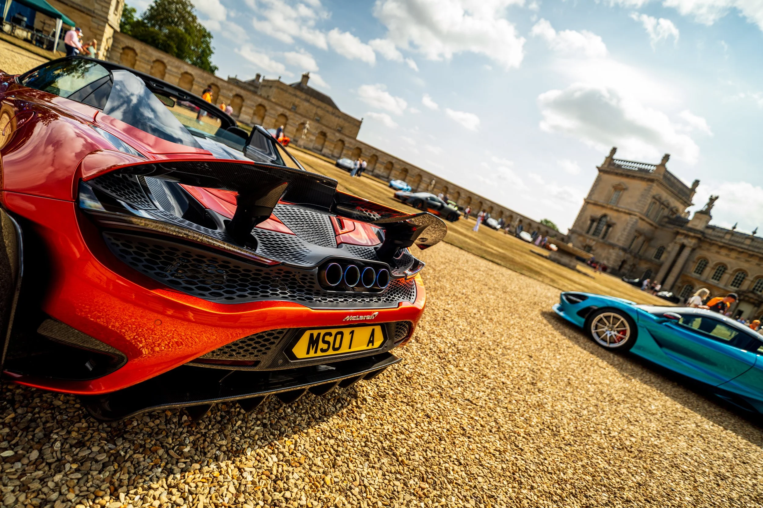 Red sports car parked on gravel with other luxury cars and historic building in background on a sunny day.
