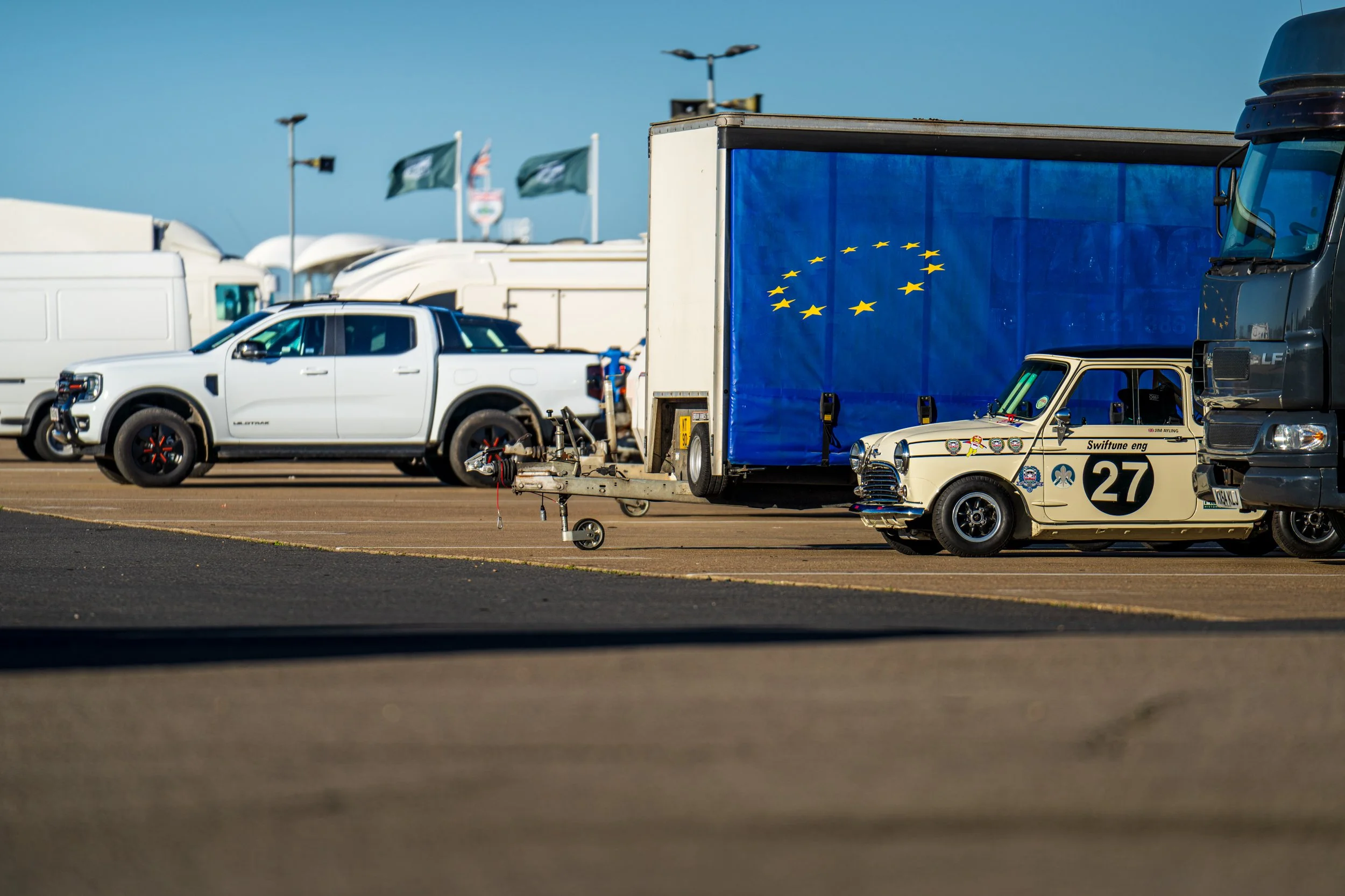 Cream Mini racing car number 27 parked among transporters in the Silverstone paddock.
