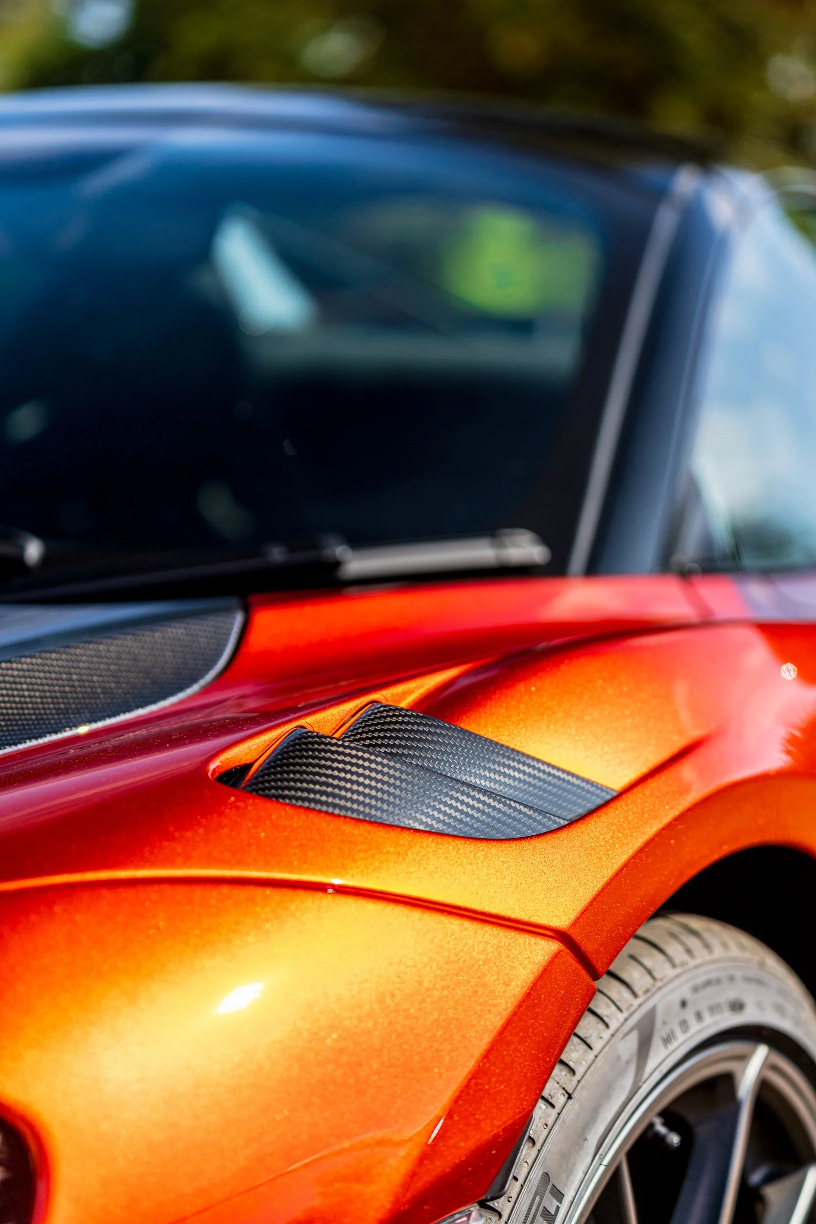 Close-up of a vibrant orange sports car with black accents and racing stripes, including a portion of the tire and the side mirror.