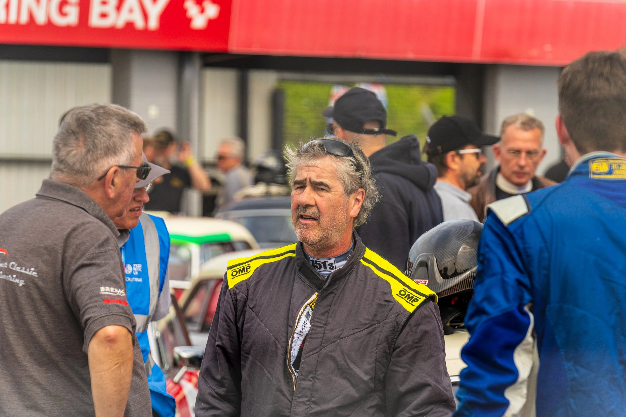Racing driver in a black suit with yellow trim in the Donington Historic Festival paddock.