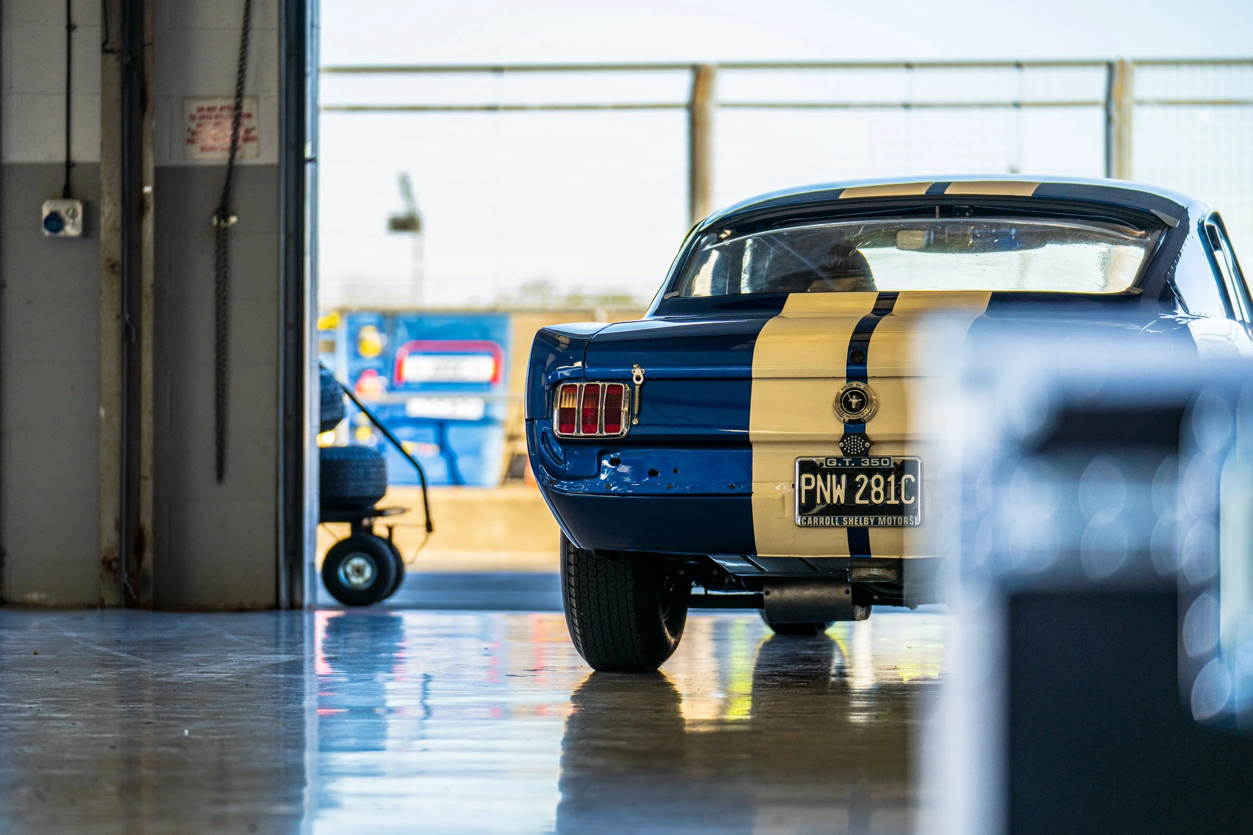Rear of a blue racing car with yellow stripes inside a Silverstone pit garage.