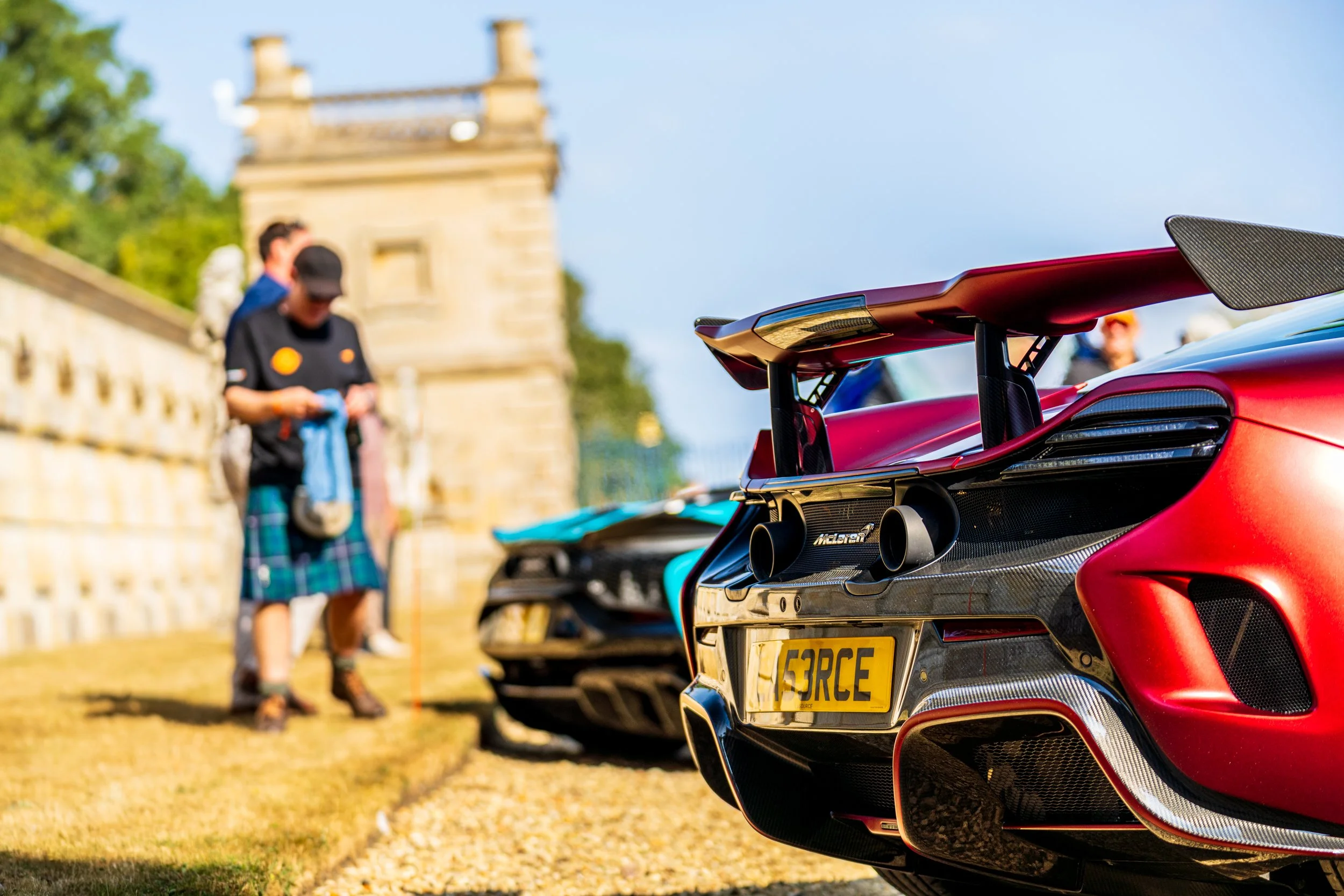 Close-up of the rear of a red sports car with a large rear wing, parked outdoors during the daytime. In the background, blurred people are standing near a stone wall, with some wearing kilts and casual clothing, and a historic building is visible und