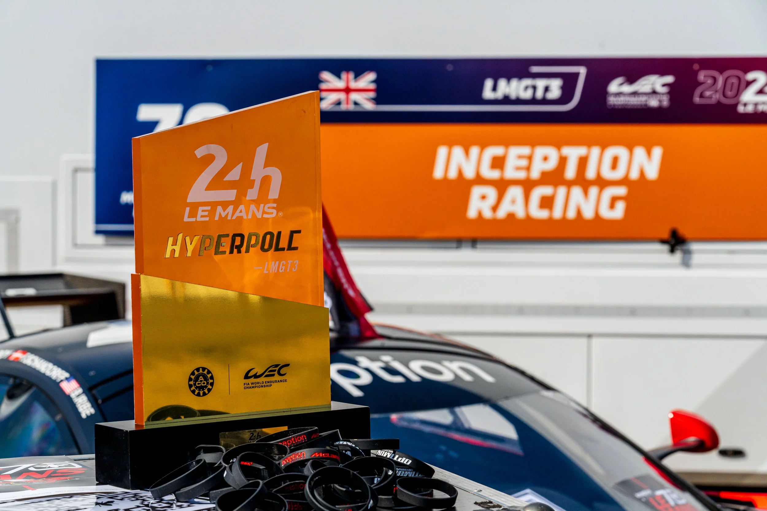 Race car setup with sponsor banners, including '24h Le Mans' and 'Inception Racing', along with racing wristbands on a table.