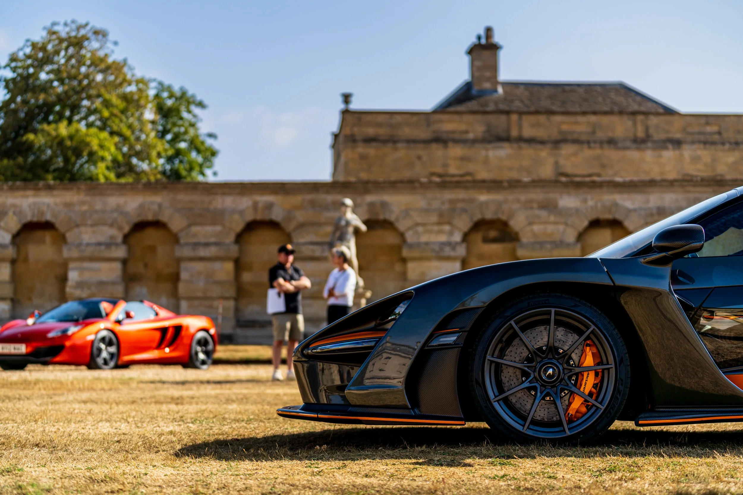 Close-up of a black sports car with orange brake calipers parked on grass, with a red sports car and two people talking in the background, and an historic stone building with an arched facade and a statue in front.