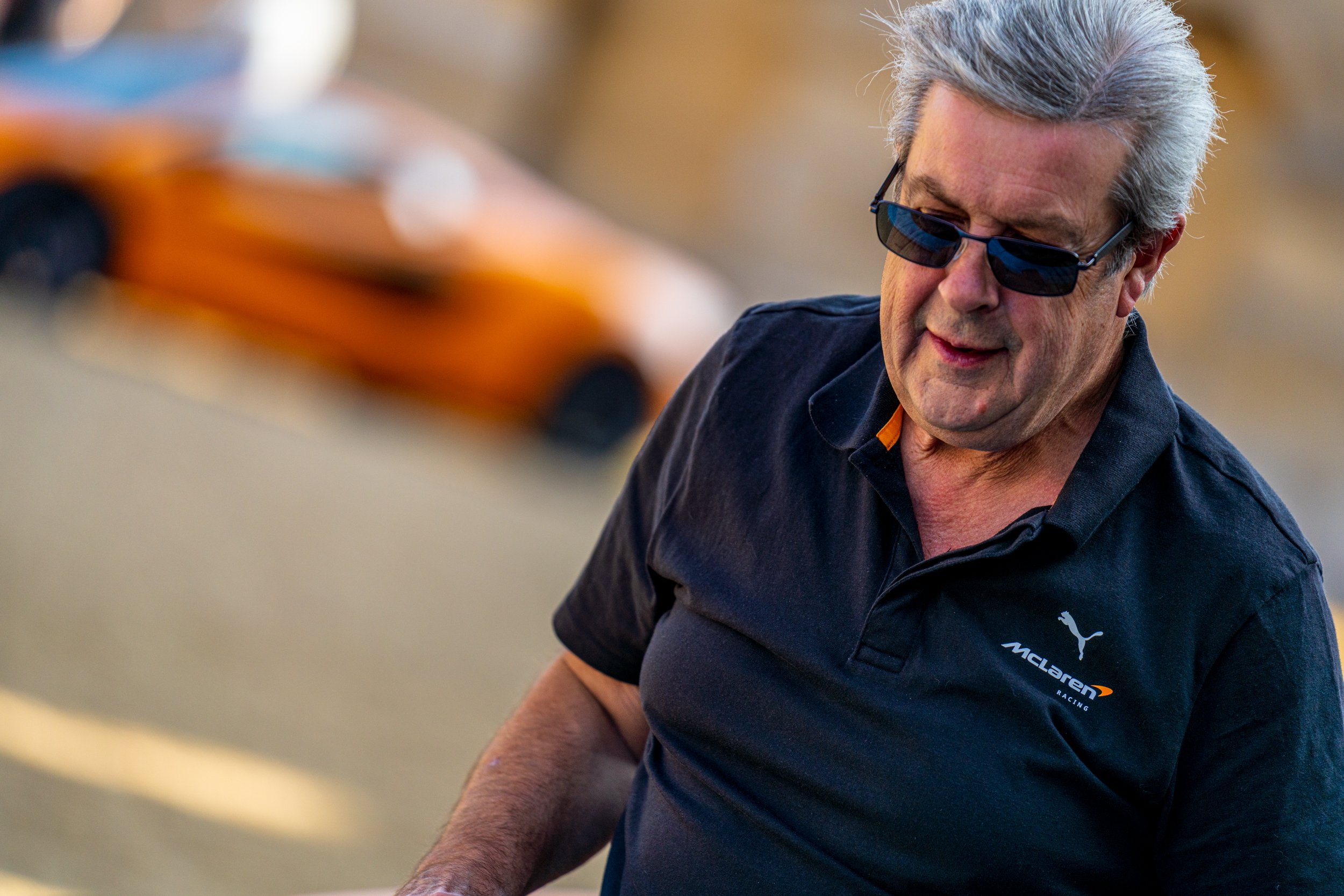 A man with gray hair wearing sunglasses and a black McLaren racing shirt, smiling and looking downward.