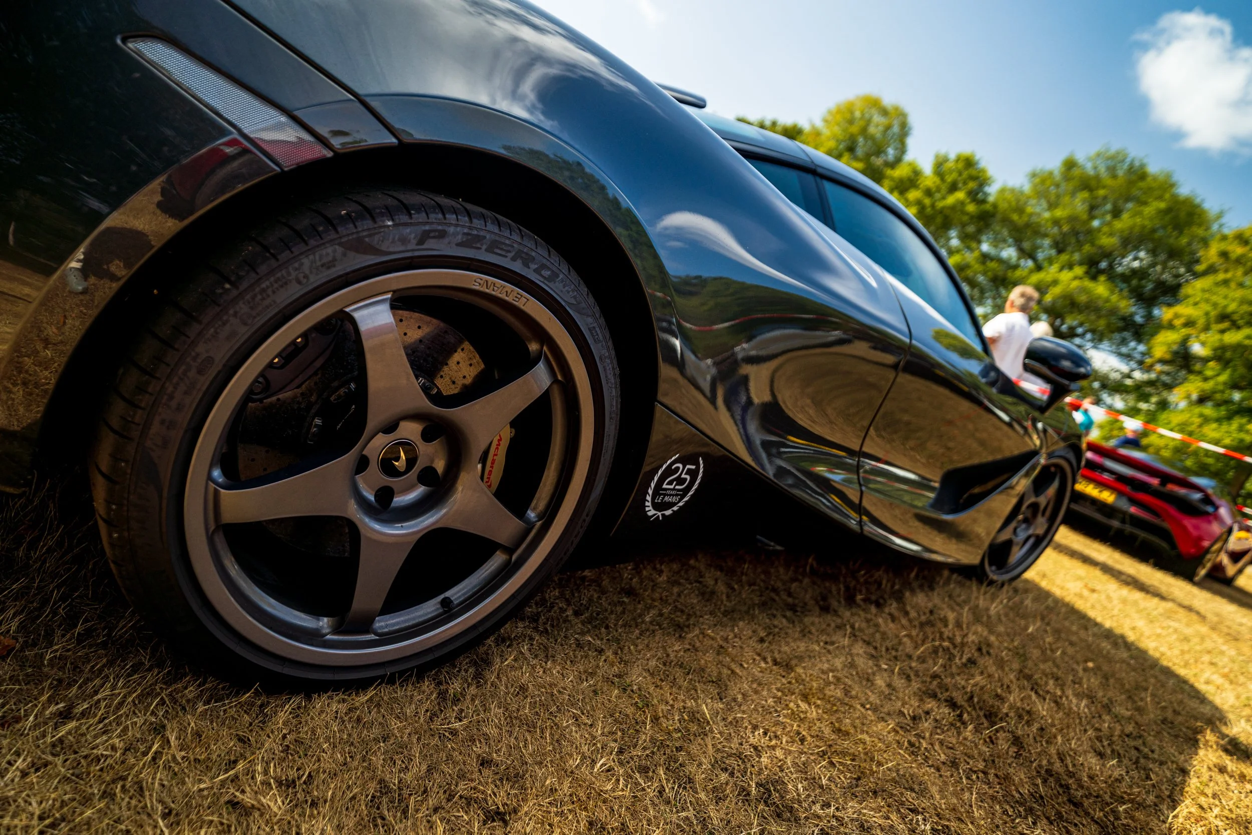 Close-up of a black sports car with a low angle view, showing the front wheel and part of the side, parked on grass with trees and a bright blue sky in the background.