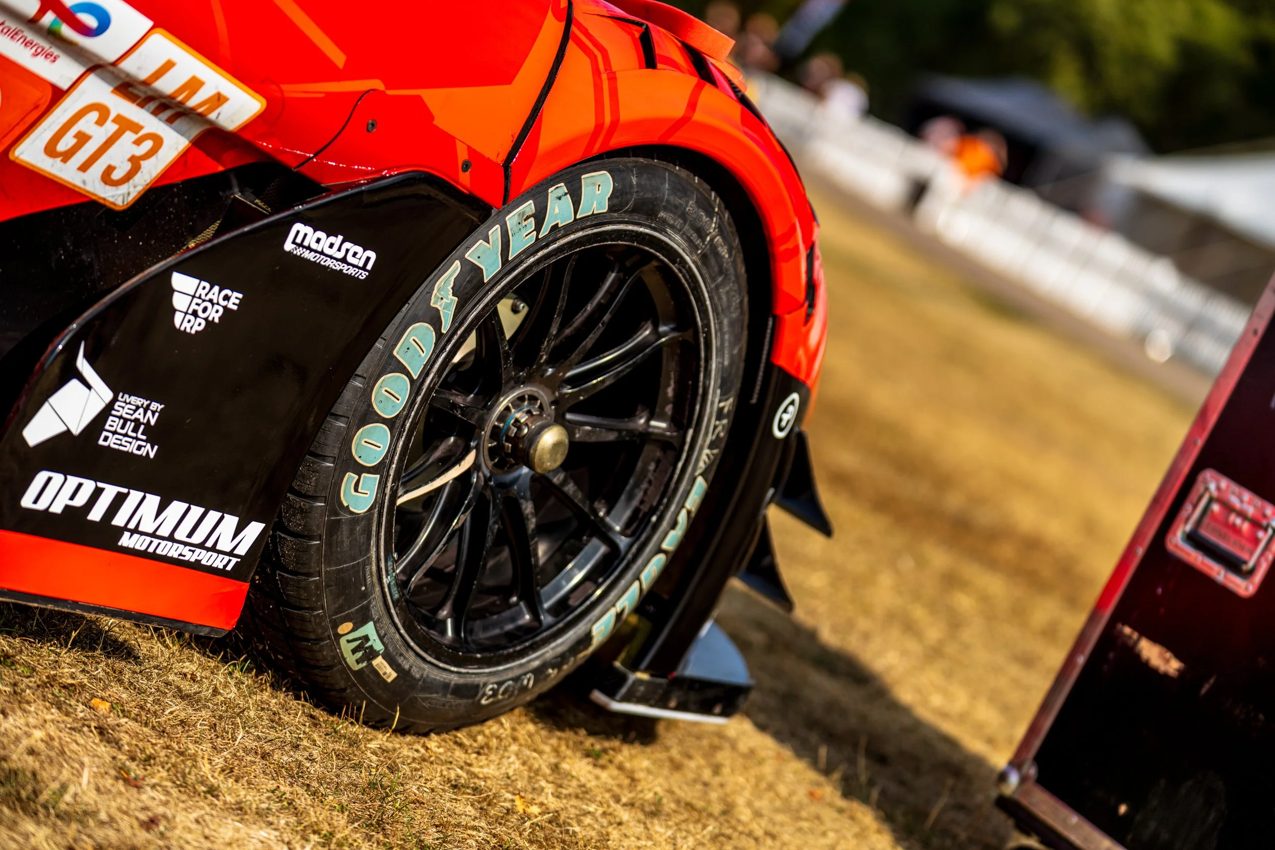 Close-up of a red race car tire with sponsorship decals, on a dirt surface with blurred spectators and equipment in the background.