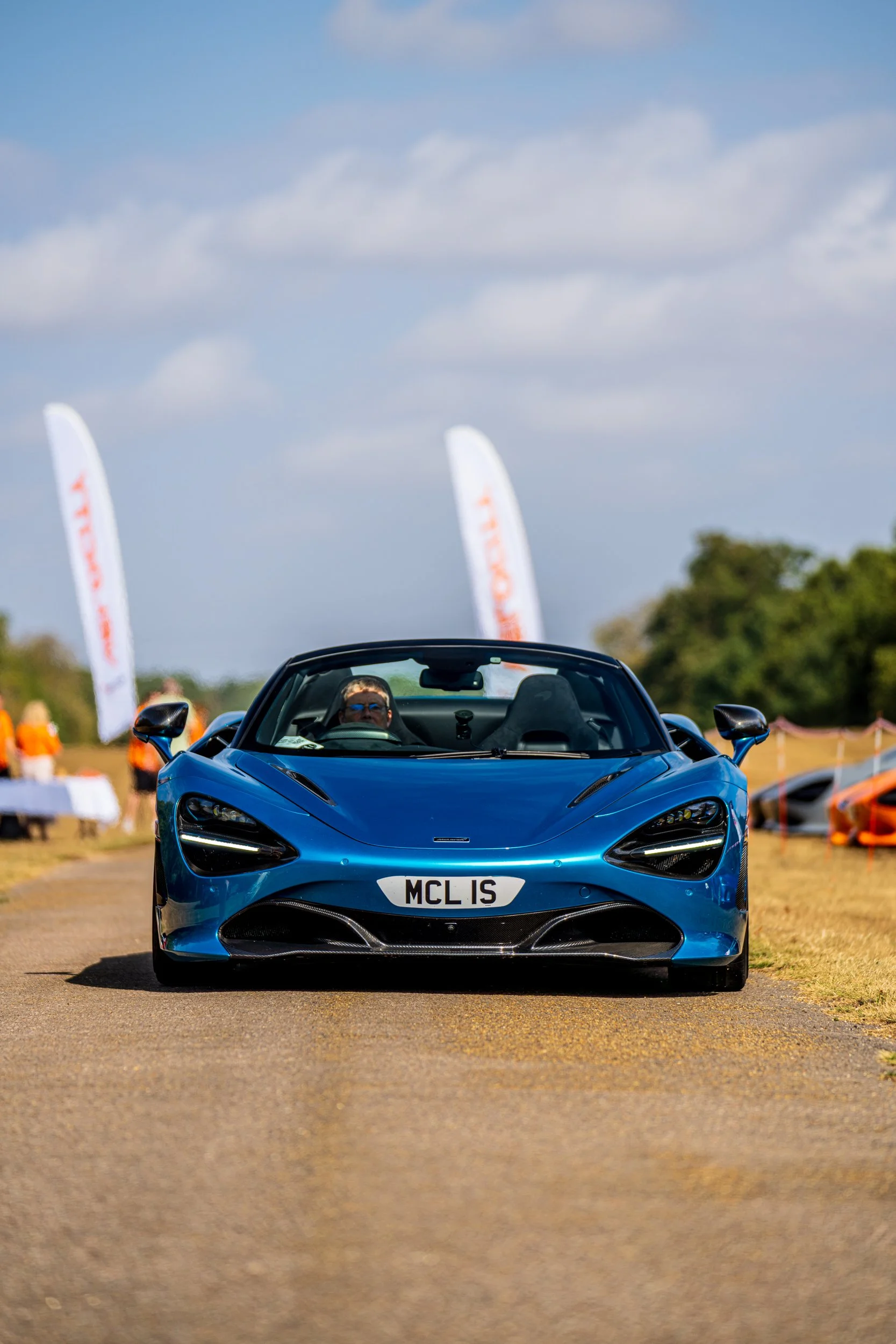 Blue sports car with a person inside, driving on a paved path. The car has a license plate that reads 'MCL 1S.' In the background, there are flags with the word 'TRIUMPH' and a group of people, some wearing orange shirts. The sky is partly cloudy.