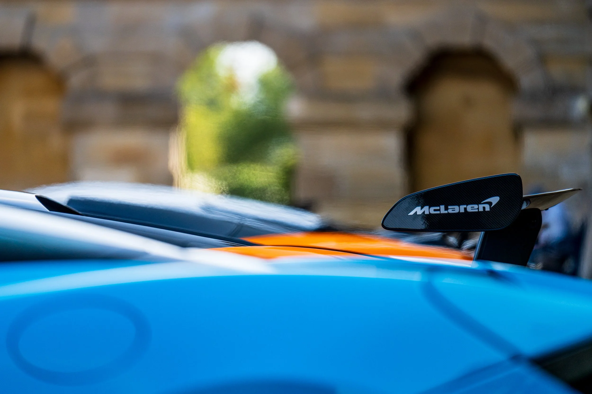 Close-up of a McLaren racing car, focusing on the rear wing with the McLaren logo, with blurred background of a tree and out-of-focus elements.