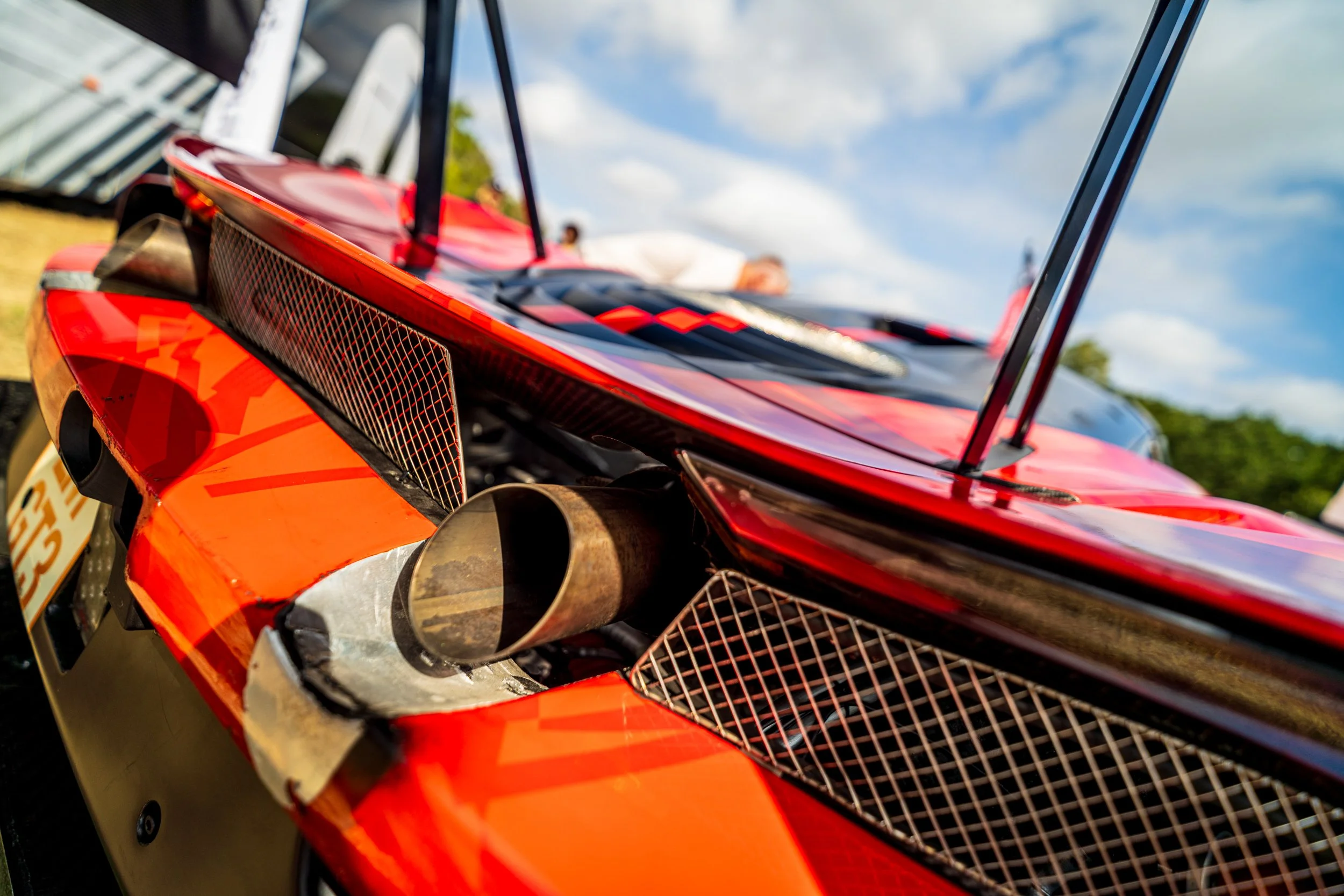 Close-up of a red race car with a focus on the front bumper, exhaust pipe, and front grille, set outdoors on a sunny day with a blue sky and clouds.