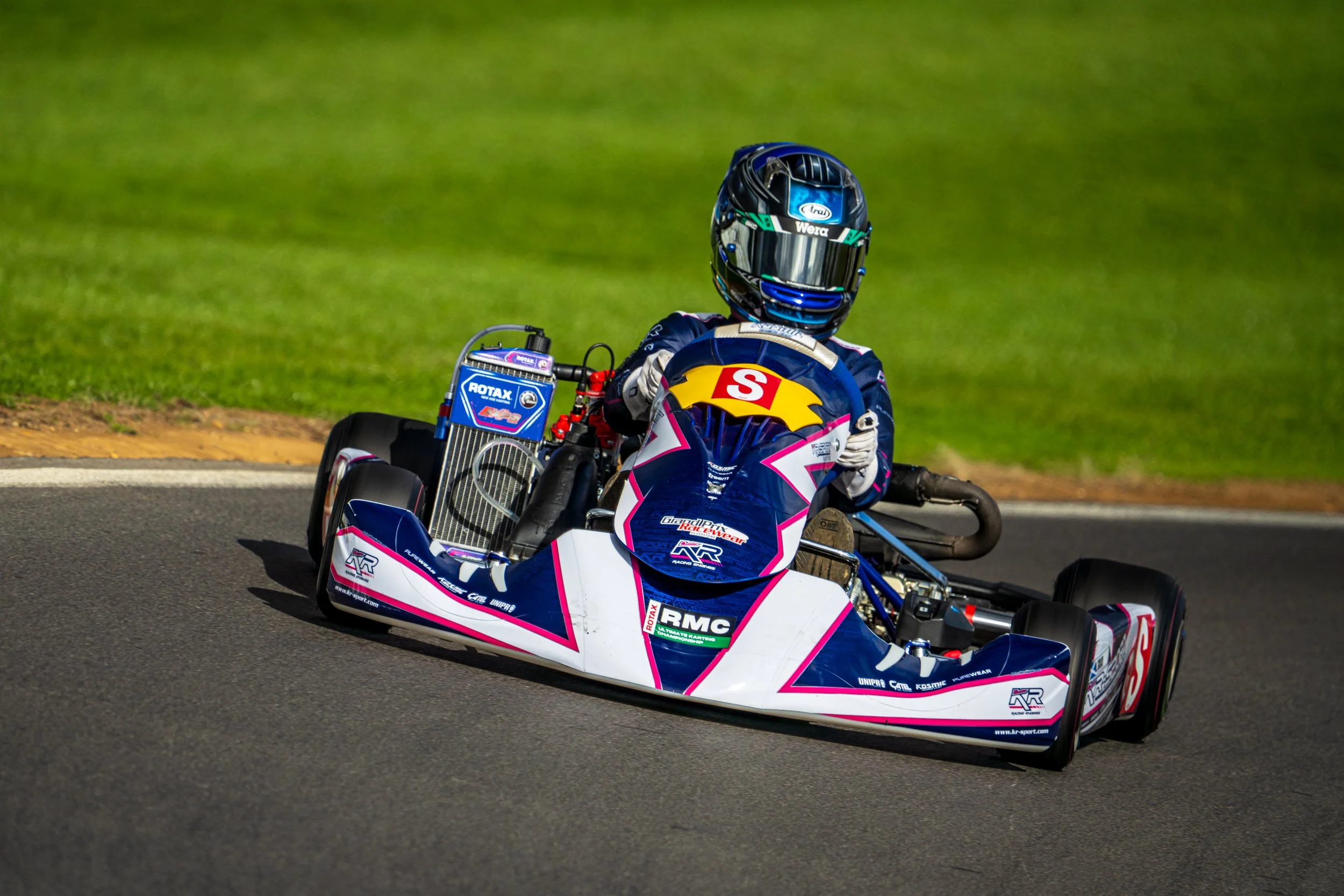Helmeted kart racer in a dark suit taking a corner at Silverstone.