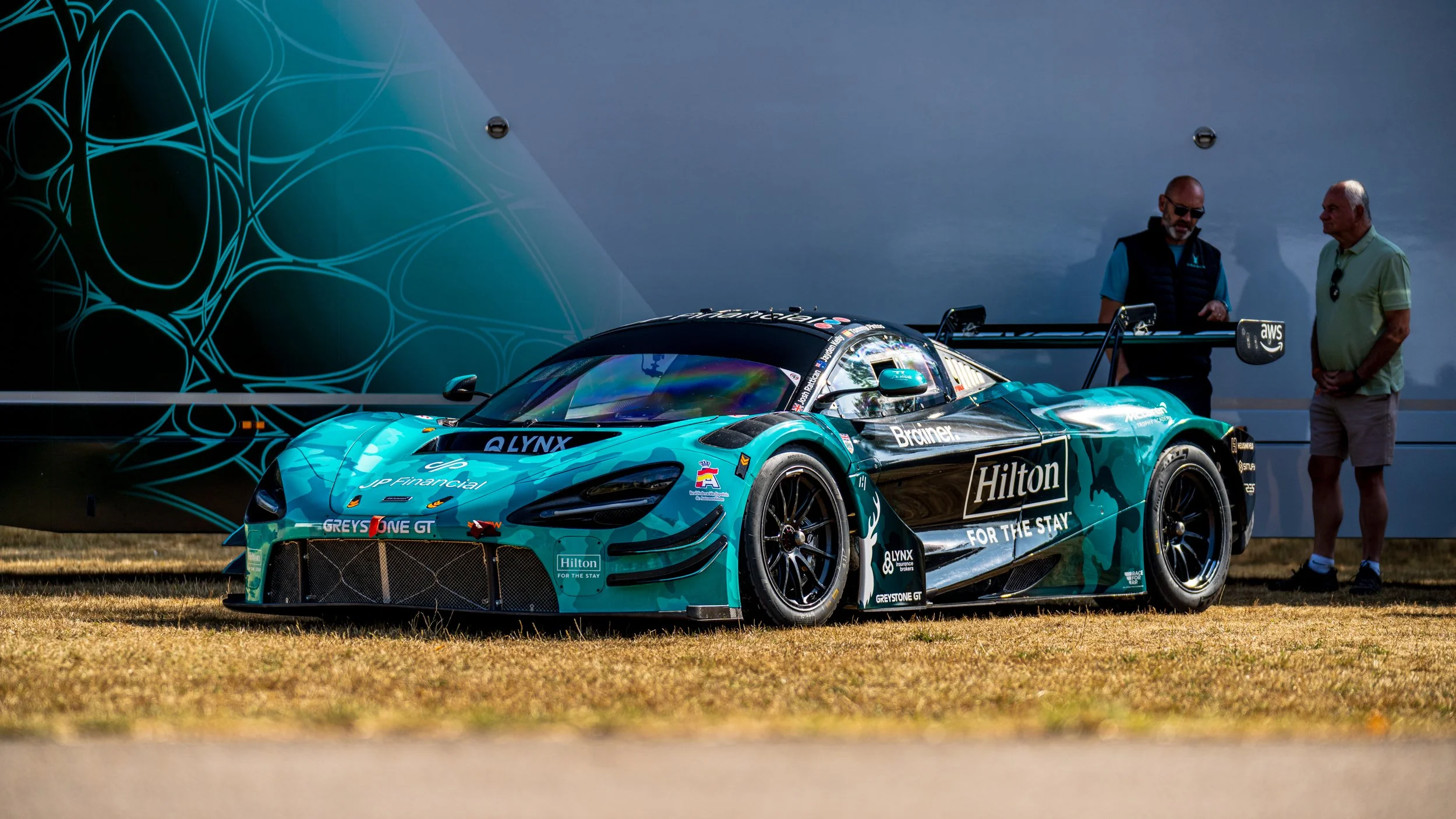 A turquoise race car with various sponsor logos, parked on grass, with two men standing nearby, one wearing sunglasses and the other with his hands in his pockets, talking.