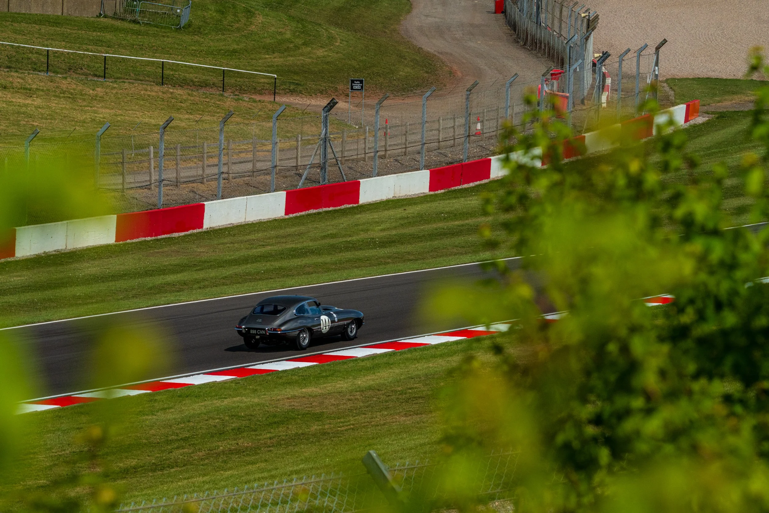 Dark racing car passing along the Donington Park circuit, framed by foliage.
