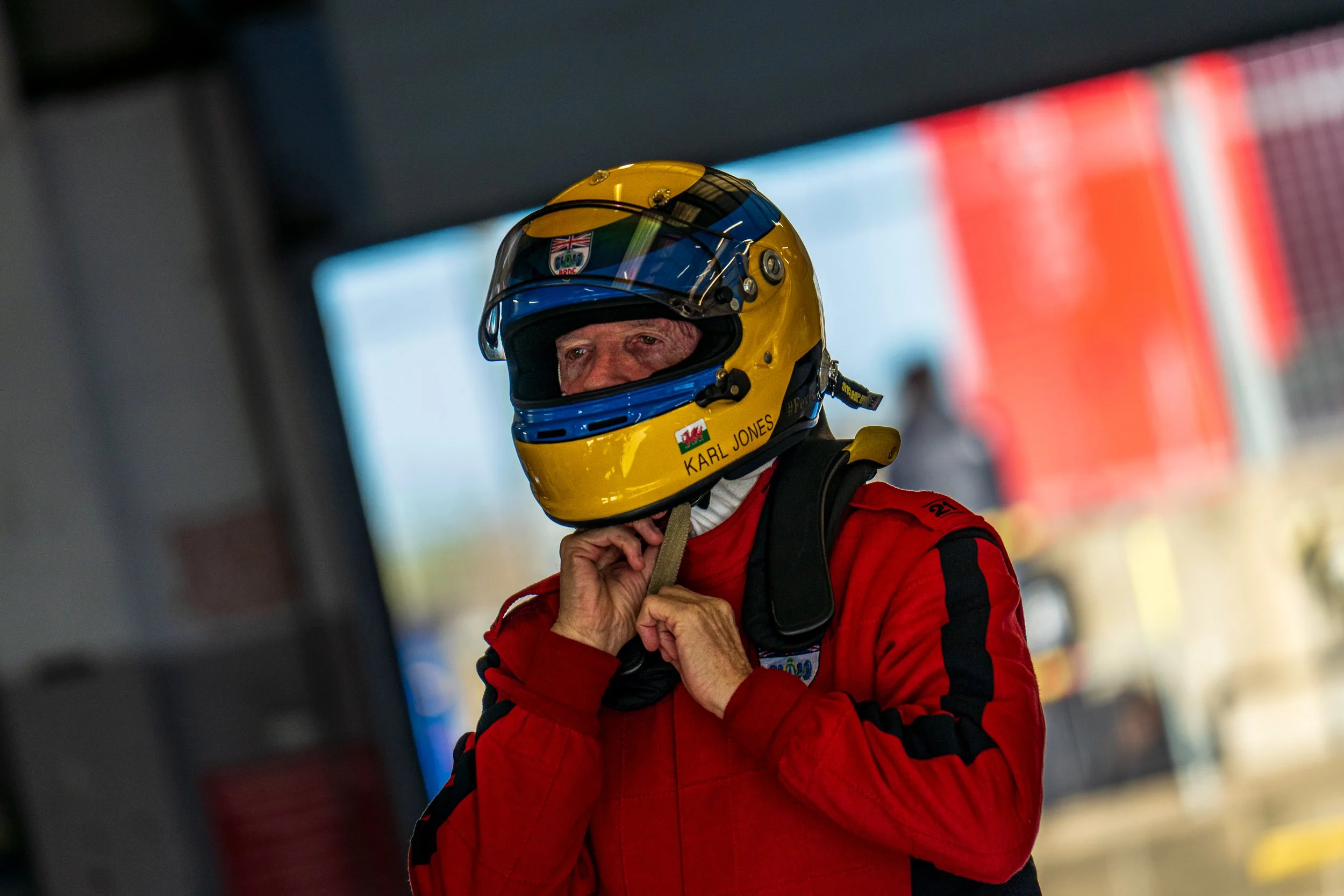 Helmeted racing driver in a red suit in the Silverstone paddock.