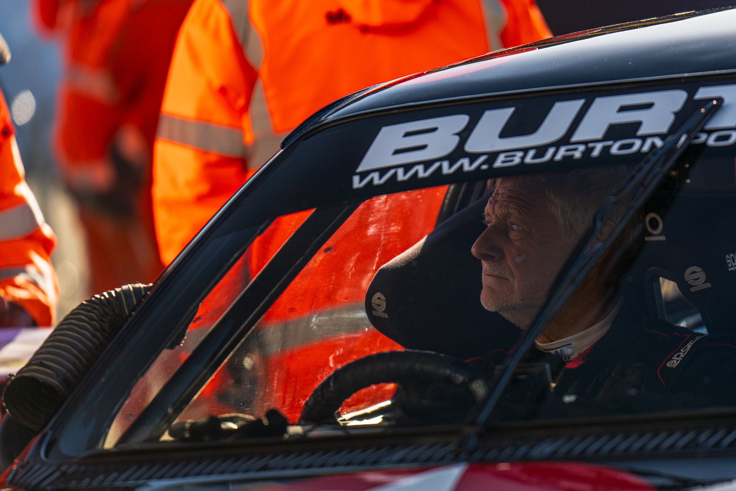 Helmeted driver seated inside a racing car beside a marshal at Silverstone.
