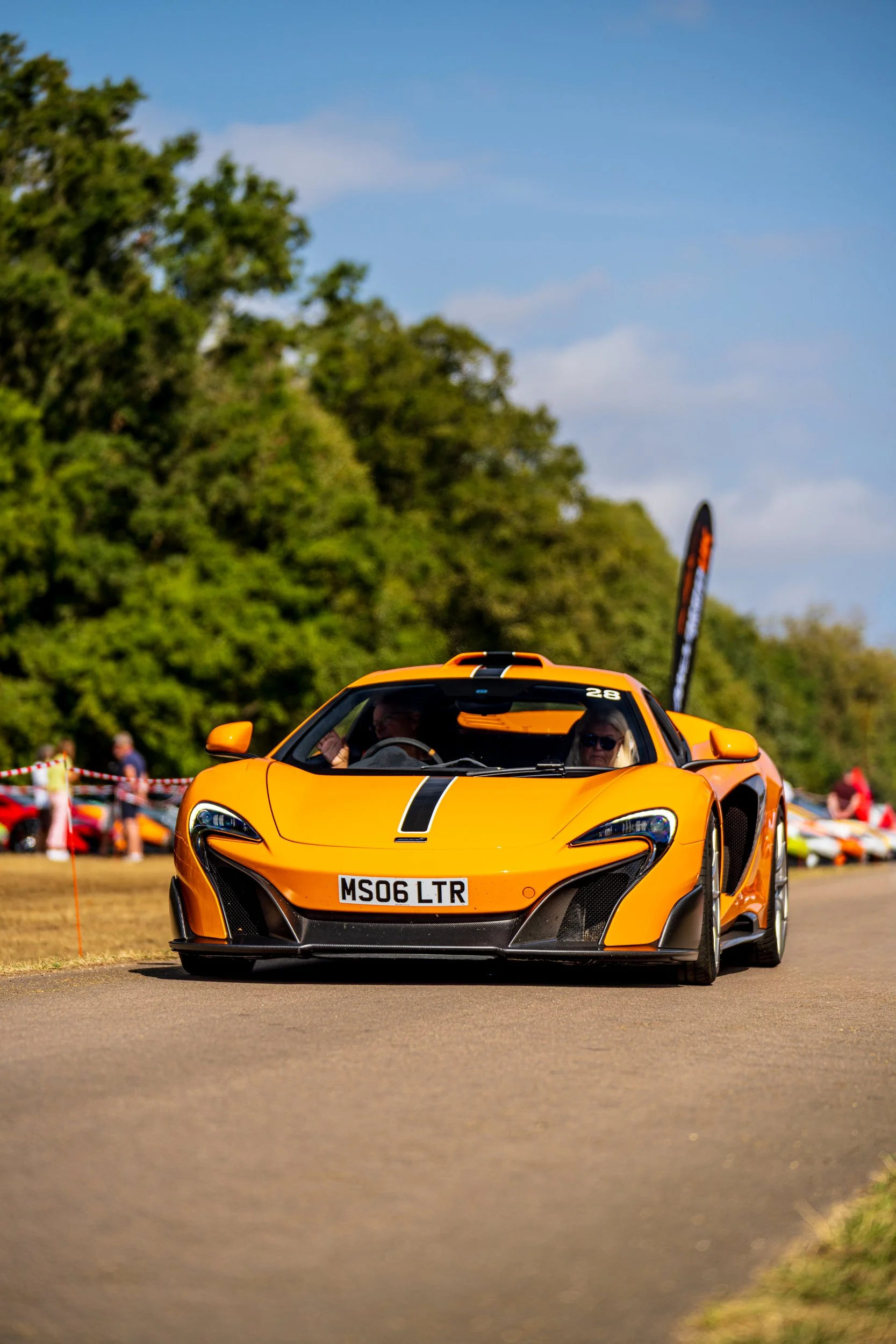 A bright orange sports car with a black stripe on the hood and the license plate MS06 LTR, driving on a paved road during an outdoor event with people and green trees in the background.