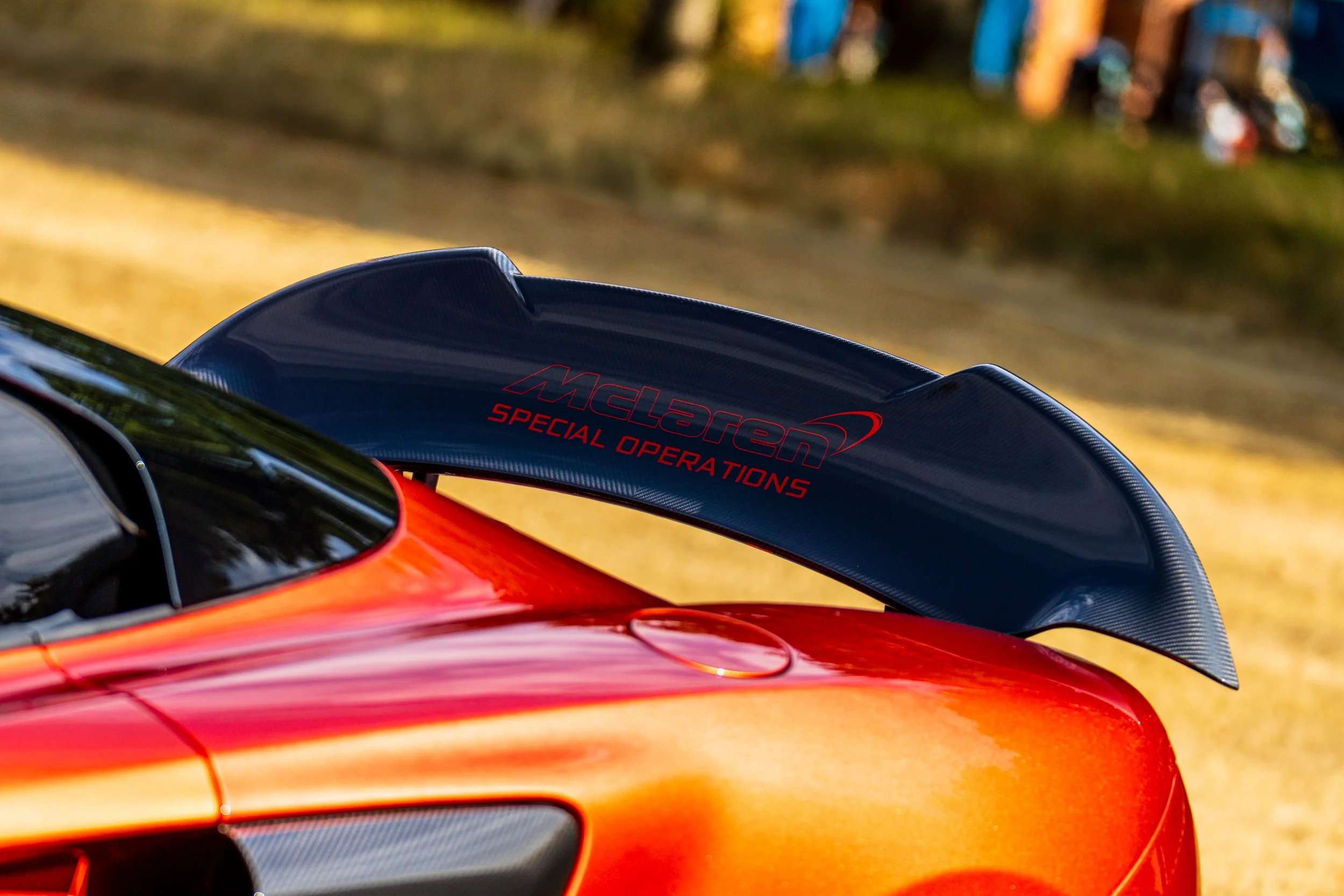 Close-up of a red race car's rear wing with the text 'McLaren Special Operations' on a black spoiler, against a blurred background of trees and colorful structures.