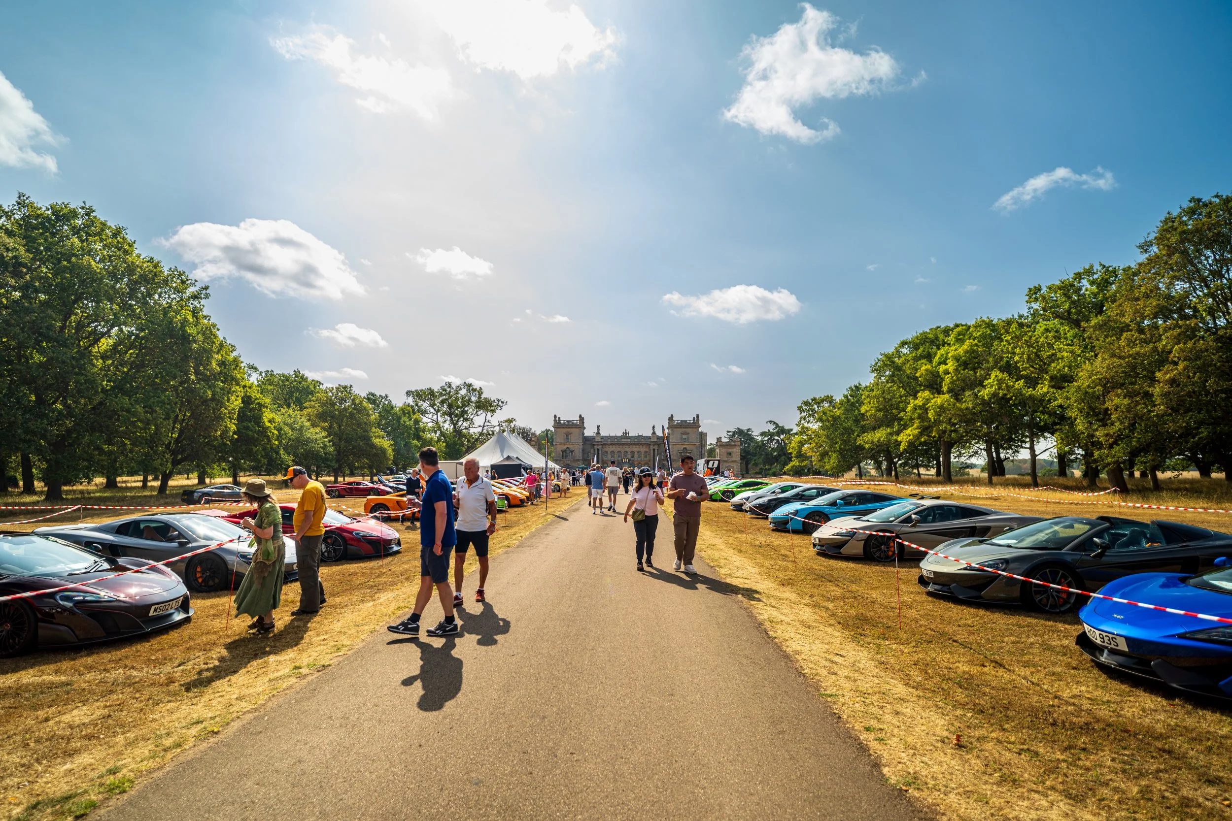 A collection of luxury sports cars parked along a pathway at an outdoor car event, with people walking and admiring the vehicles, trees lining the area, and a historic building with towers in the background on a bright sunny day.