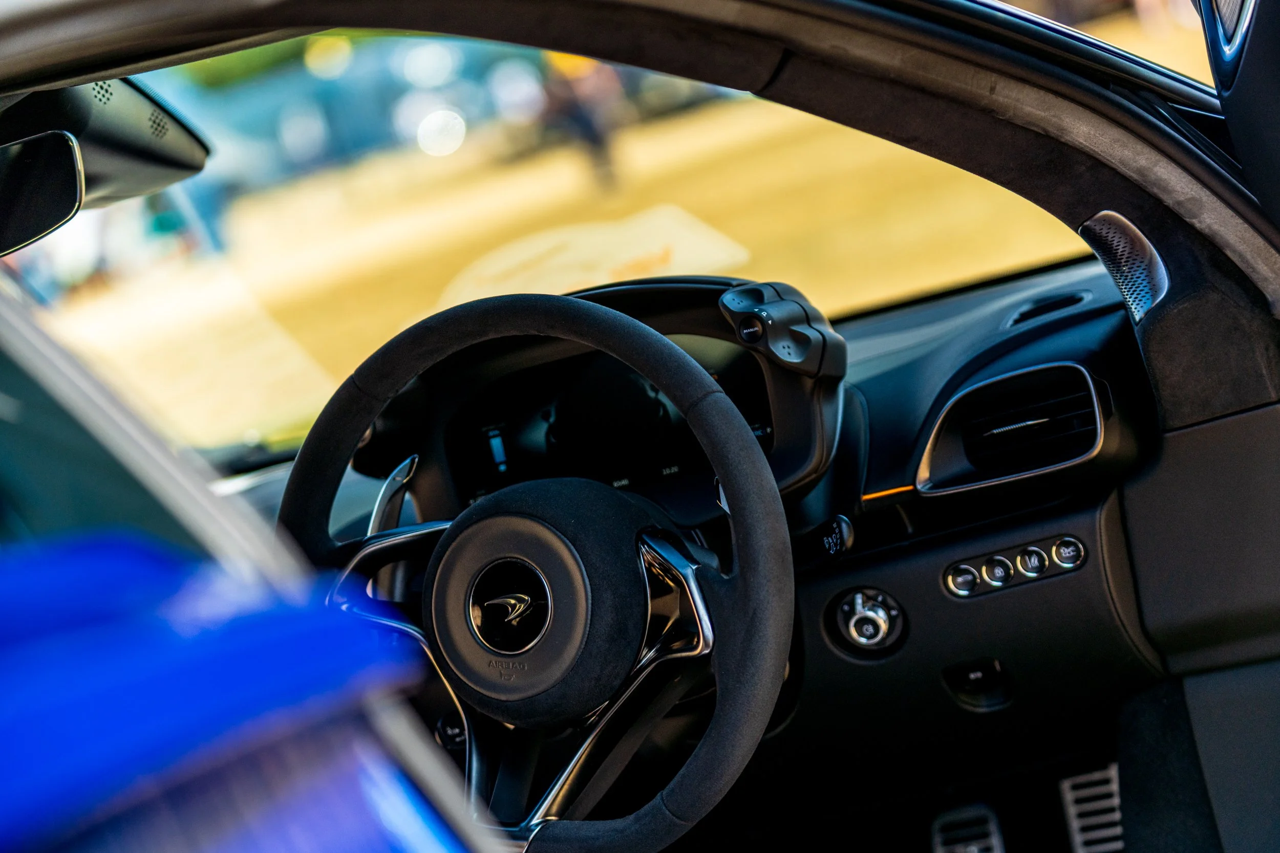 Interior view of a modern car with a steering wheel, dashboard, and control buttons, seen through the windshield with a blurred outdoor background.