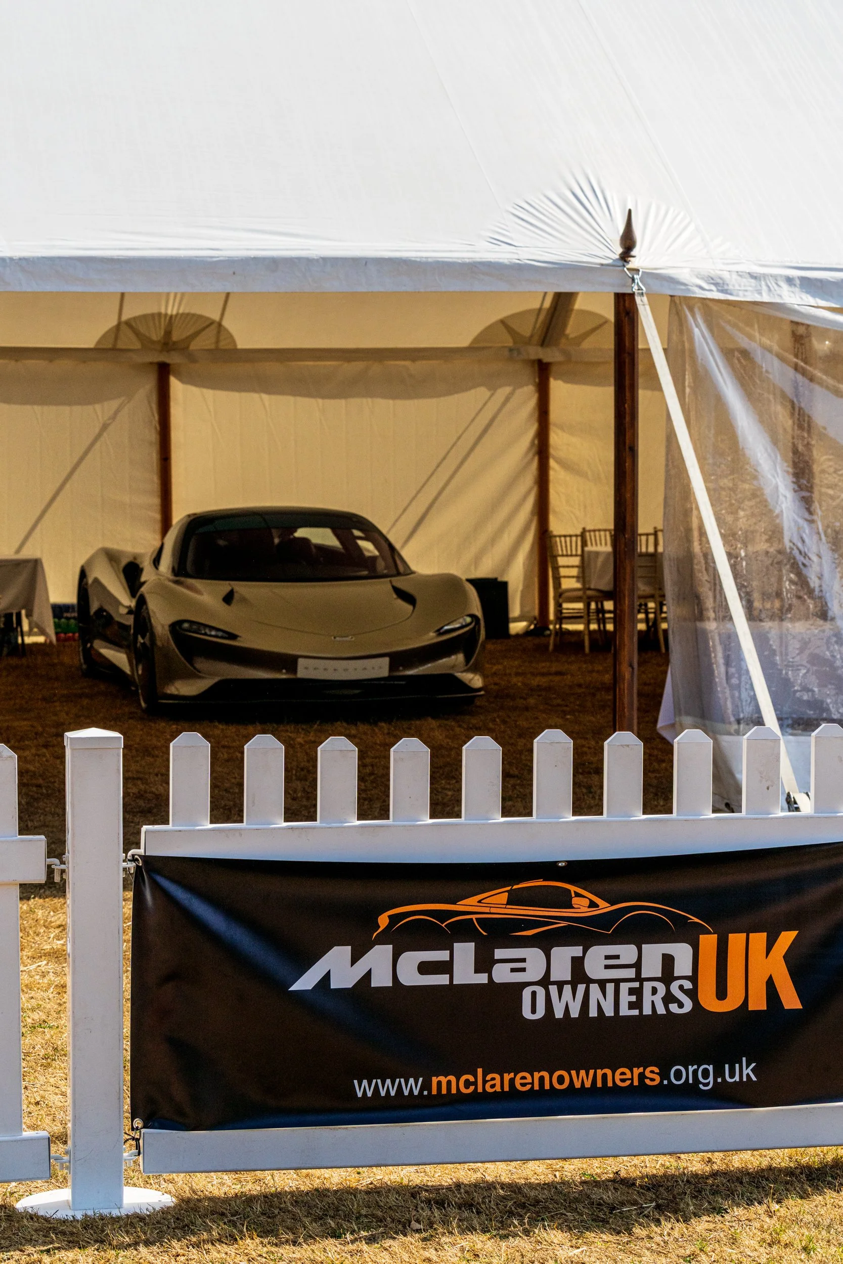 A silver McLaren sports car parked inside a white event tent with wooden posts. A black McLaren Owners UK banner hangs in front of a white picket fence in the foreground.