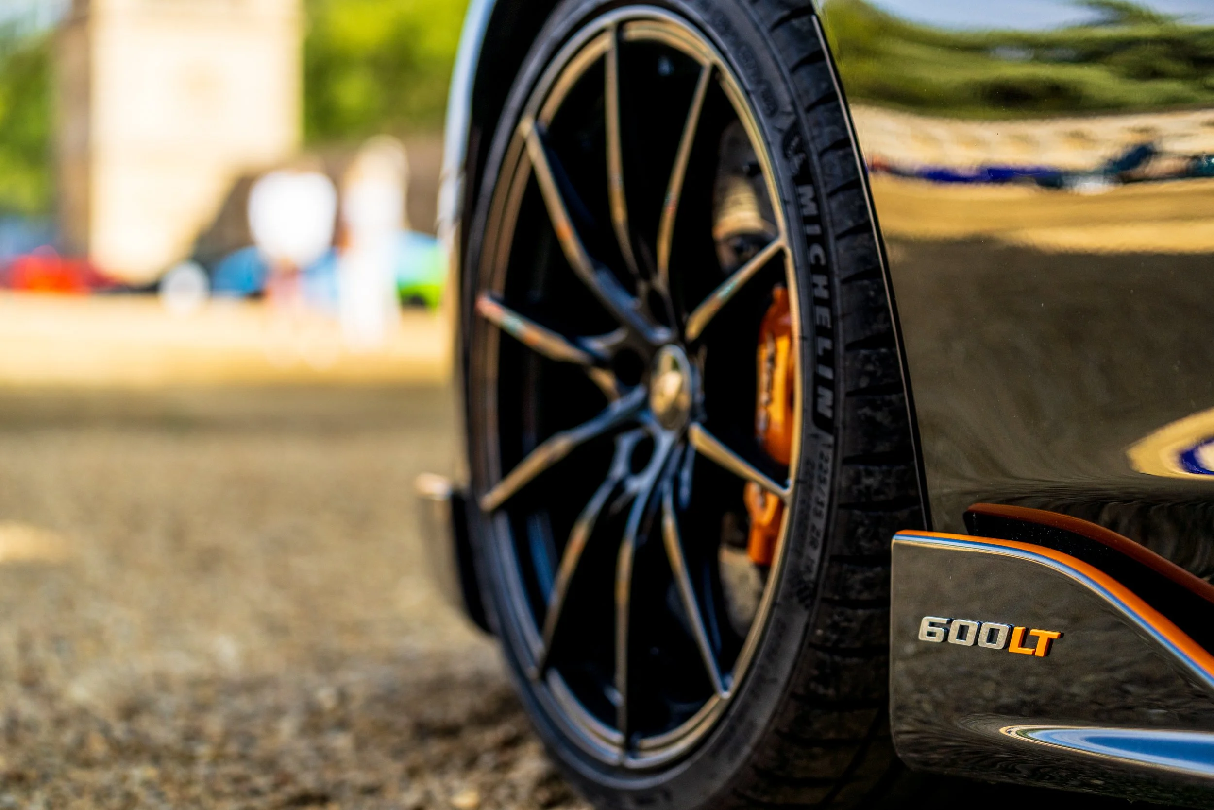 Close-up of a black luxury sports car tire with custom black and orange rims, parked on a street during daytime.