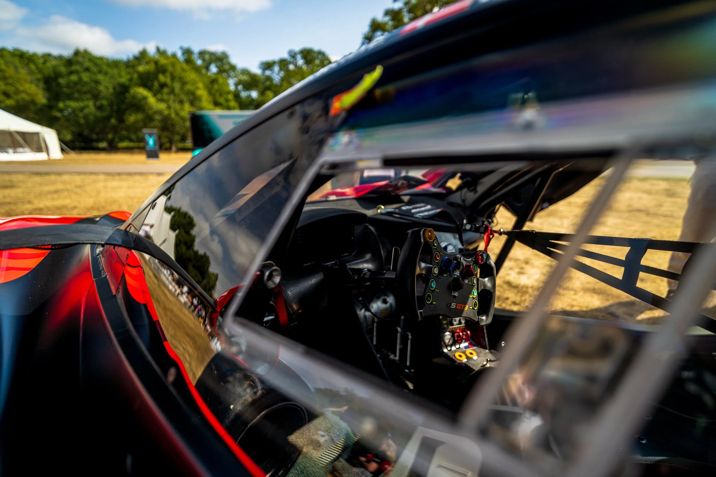 Close-up view from inside a race car cockpit, showing steering wheel with colored buttons, dashboard, and canopy with reflections, with race track and trees in the background.