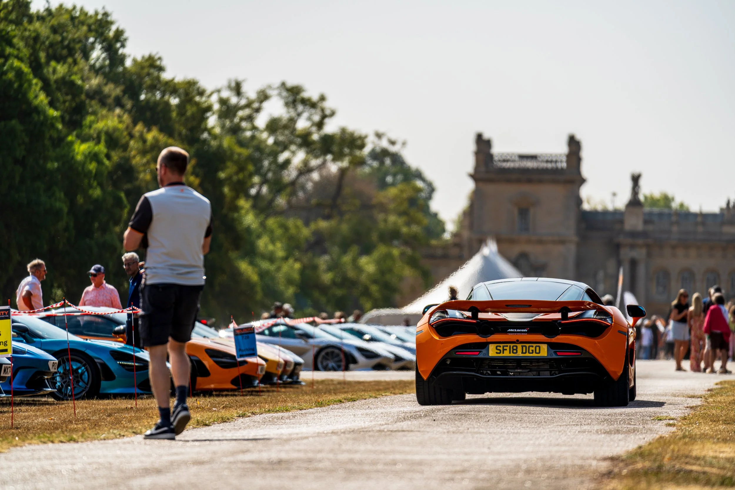 A line of luxury sports cars, including a McLaren with the license plate 'SF18 DGO', displayed outdoors at a car show with people walking around and viewing the vehicles. There is a historic building and trees in the background.