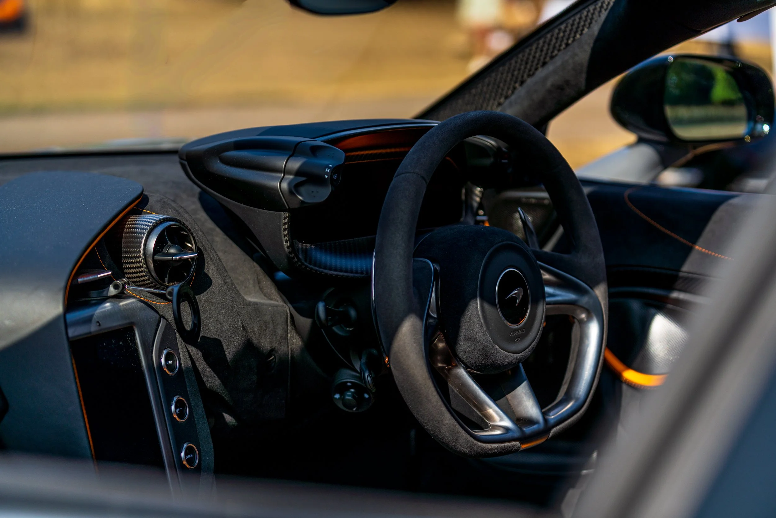Close-up view of the interior dashboard and steering wheel of a luxury sports car. The dashboard features black and orange accents, and a carbon fiber trim near the air vents. The steering wheel is wrapped in suede material.