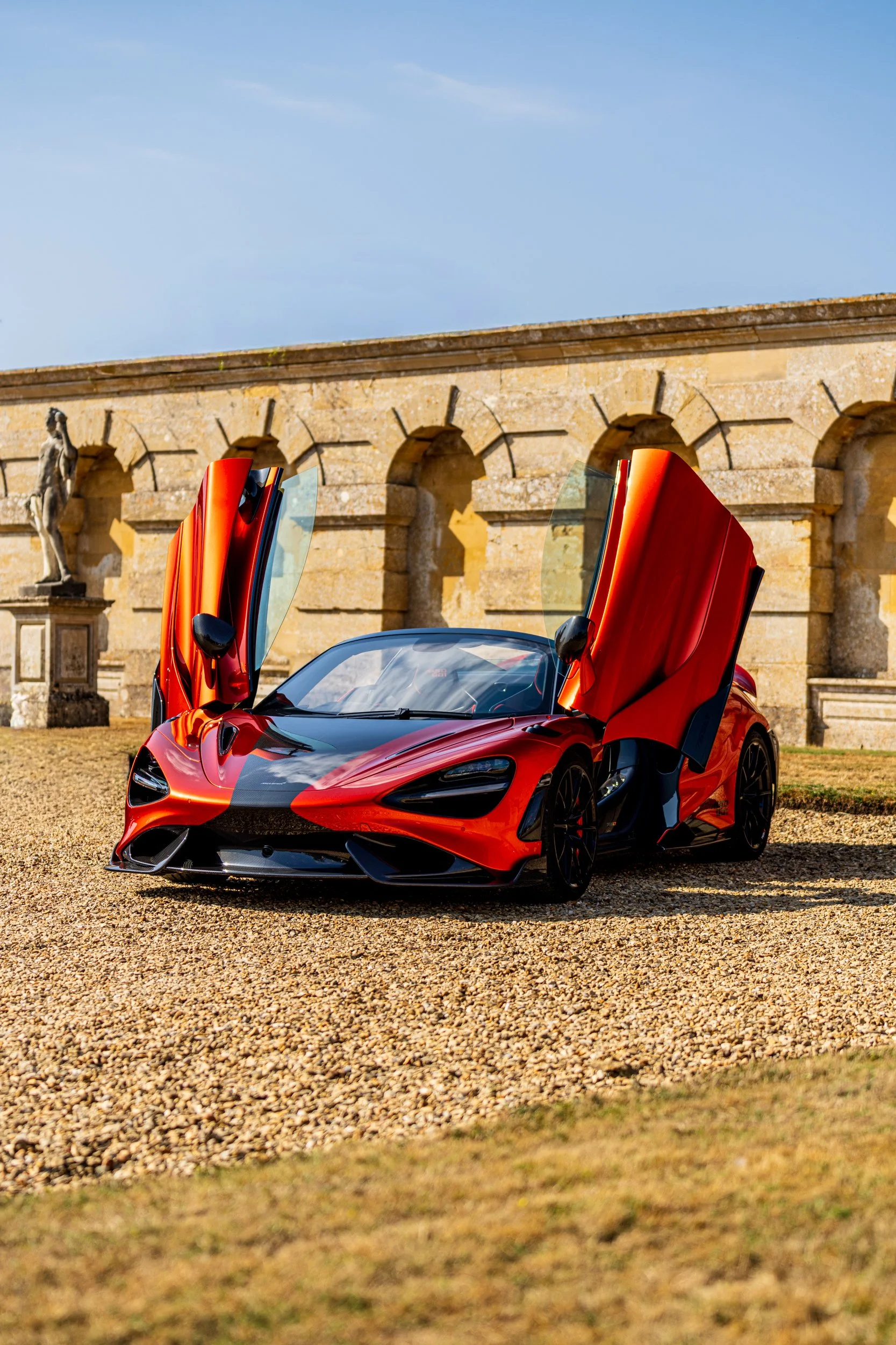 Red sports car with open butterfly doors parked on gravel in front of a historic stone building with statues and archways.