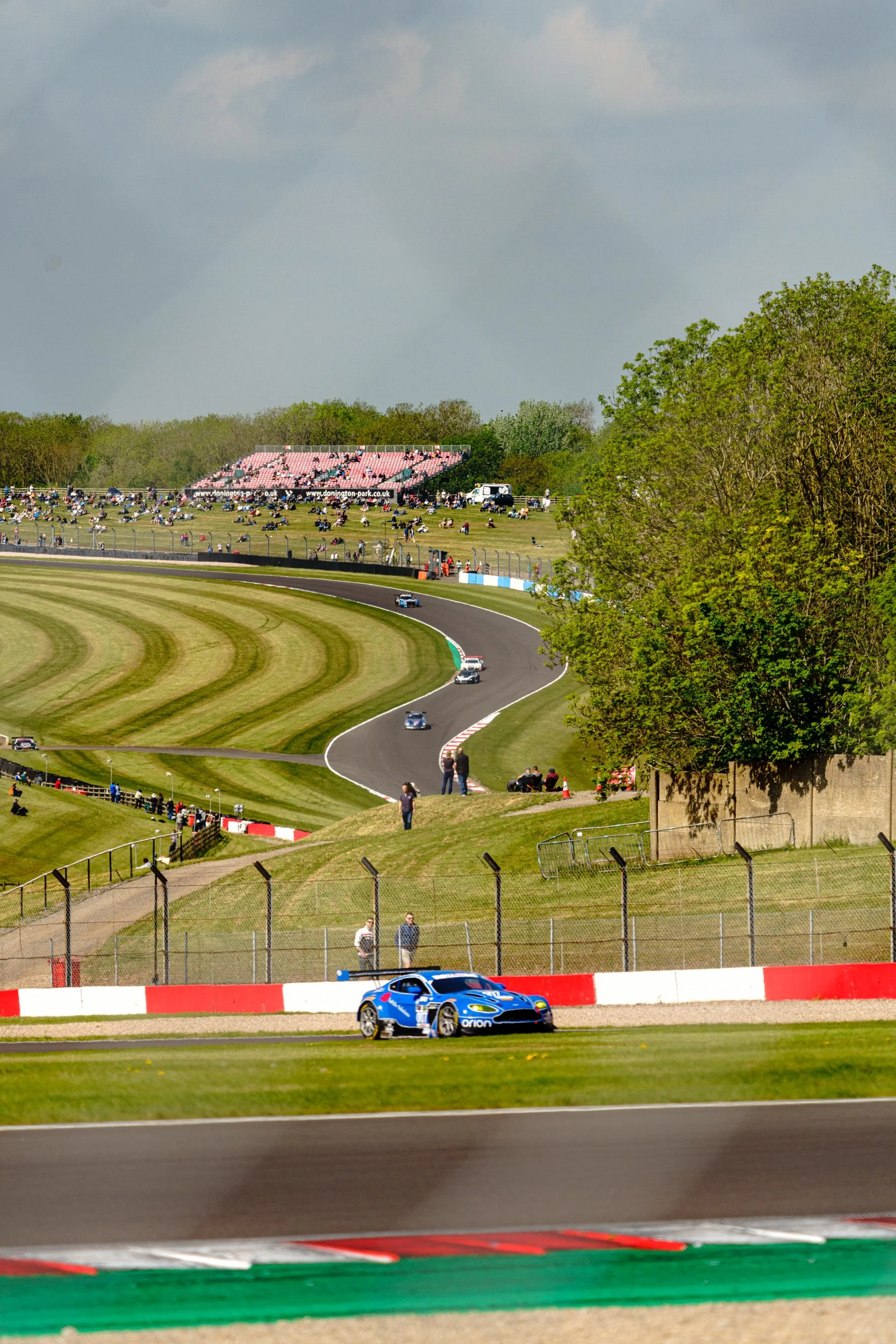 A blue racing car on the sweeping Donington Park circuit, with spectators on the hillside.