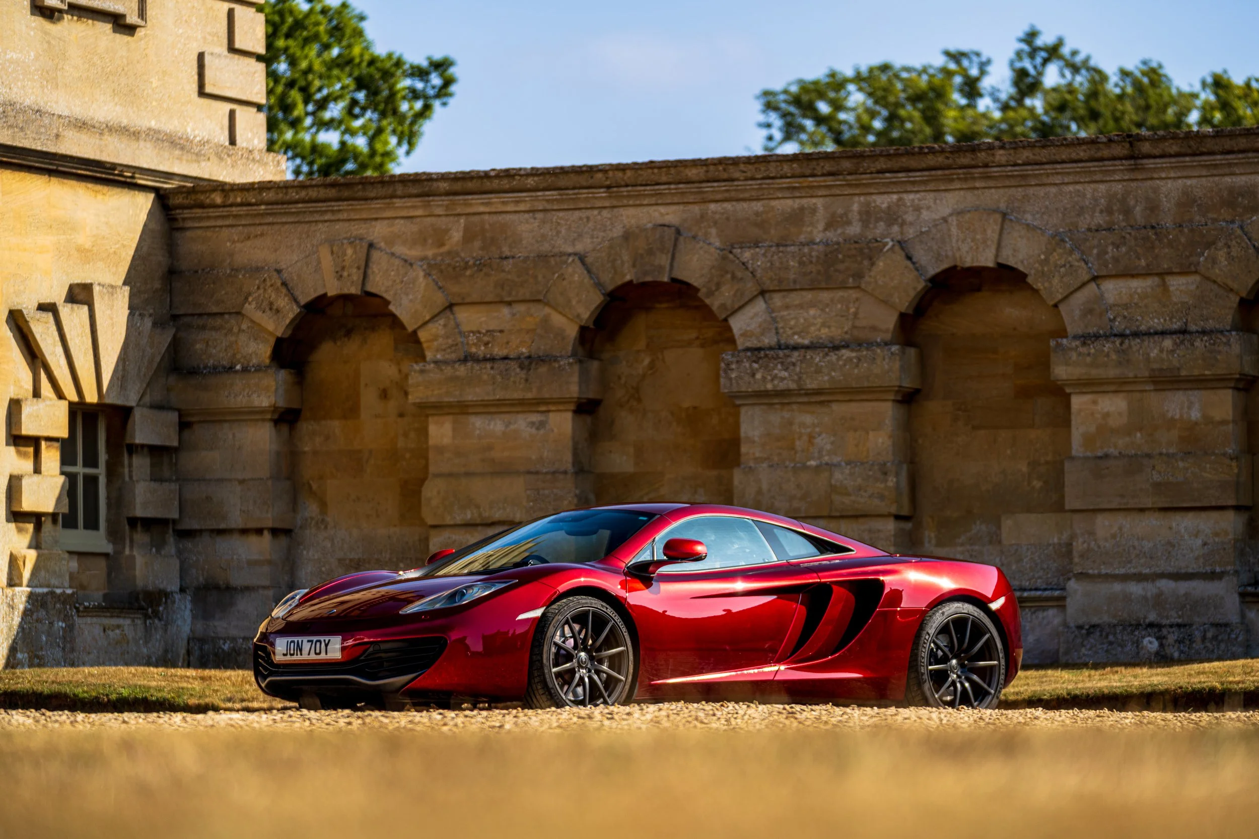 A sleek red sports car with a black roof parked on a grassy area in front of an old stone building with archways and a window. Trees and a blue sky are visible in the background.