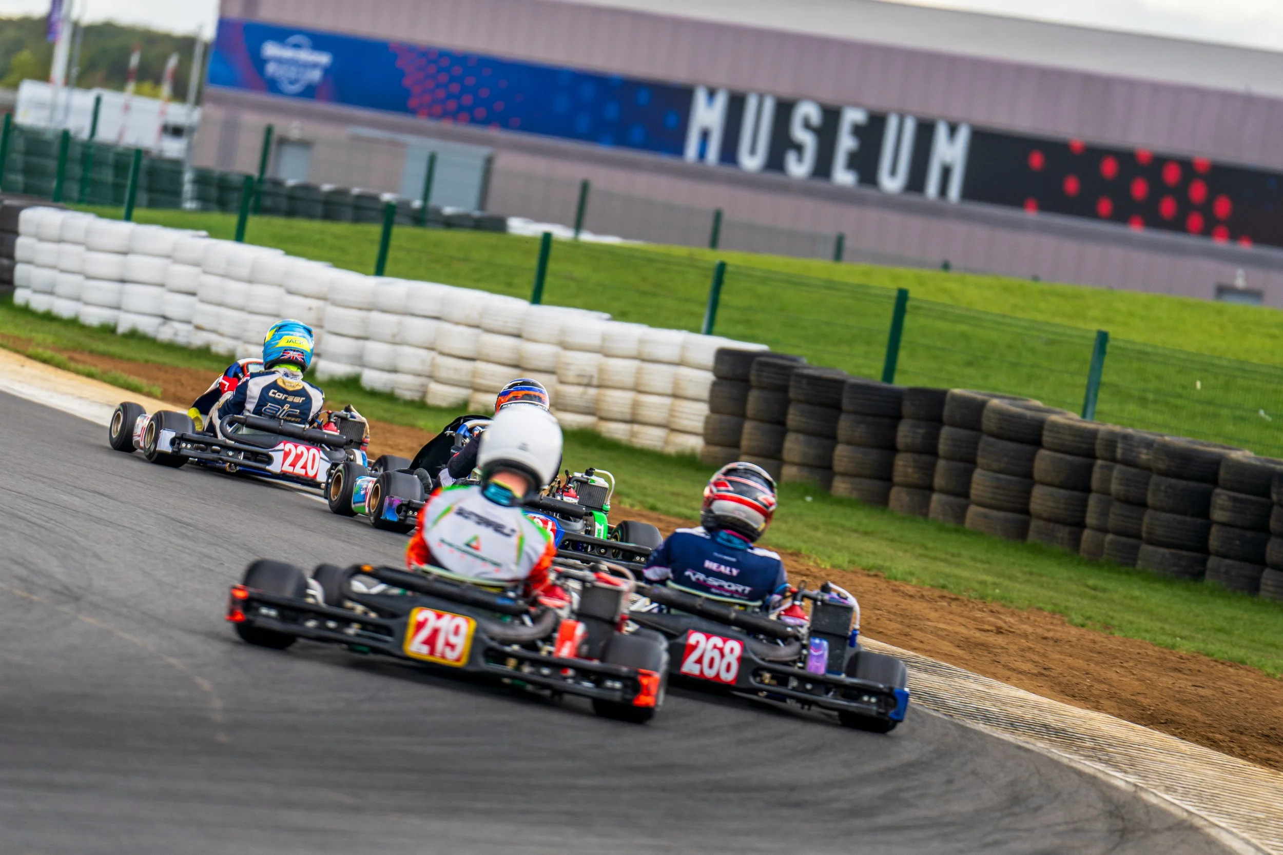 A group of kart racers competing with the Silverstone Museum building in the background.