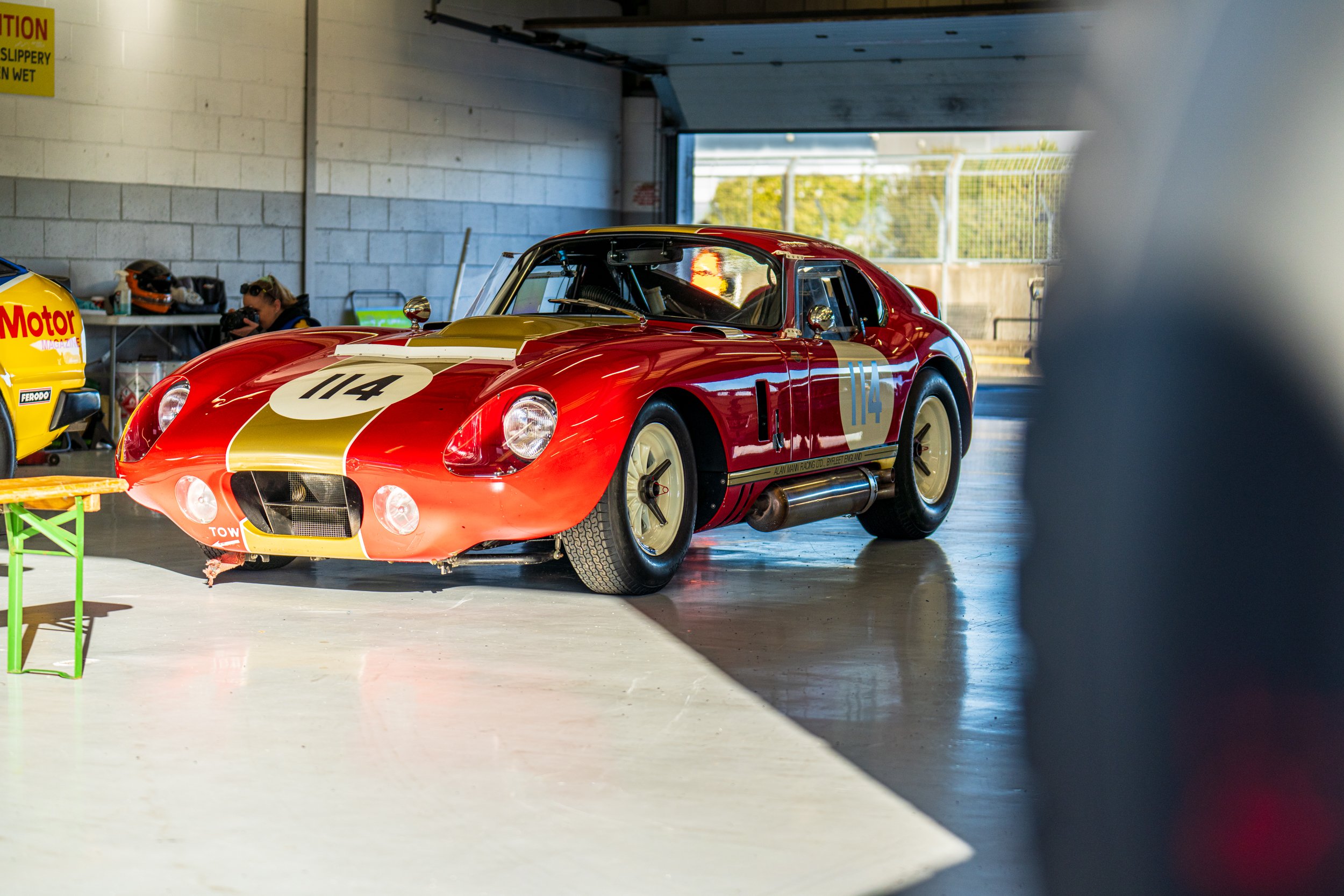 Red and white classic saloon racing car inside a Silverstone pit garage.