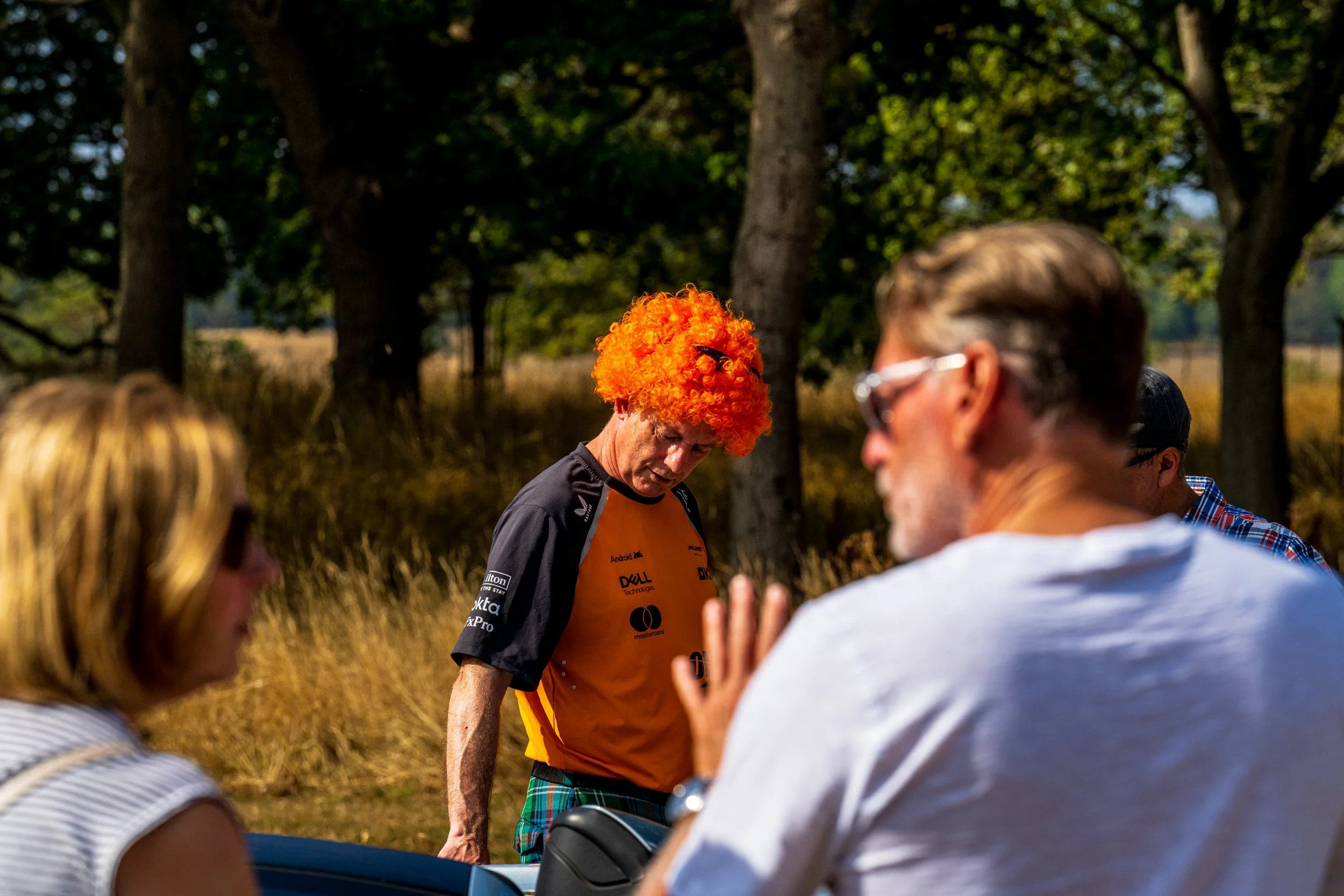 People gathered outdoors in a circle, one person wearing a bright orange curly wig and a orange Mclaren jersey, engaging in conversation, with trees and a field in the background.