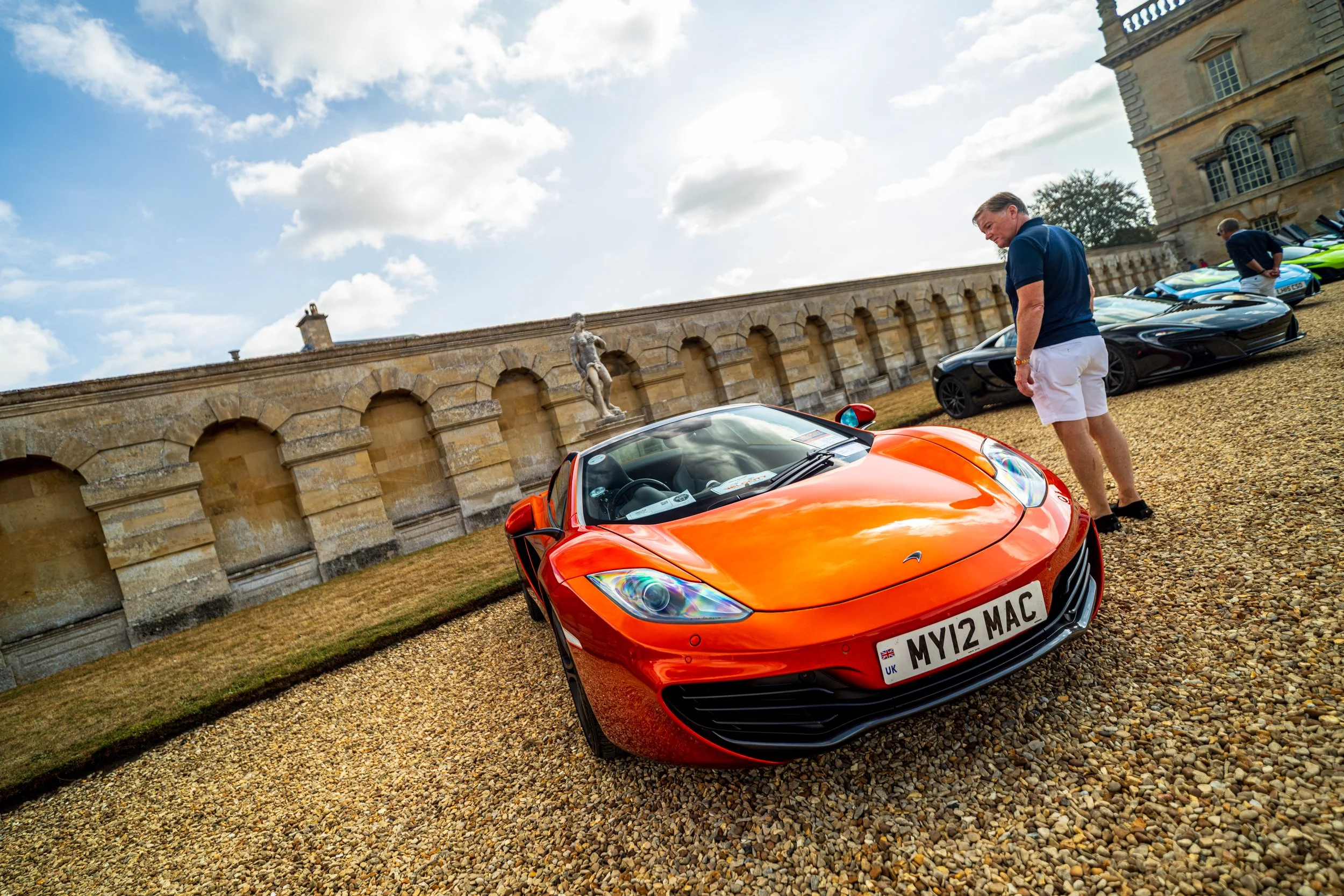 A bright orange sports car with a UK license plate parked on a gravel driveway at a historic estate. A man wearing a blue shirt and white shorts is standing nearby, looking at the car. Several other luxury sports cars are parked in the background, an