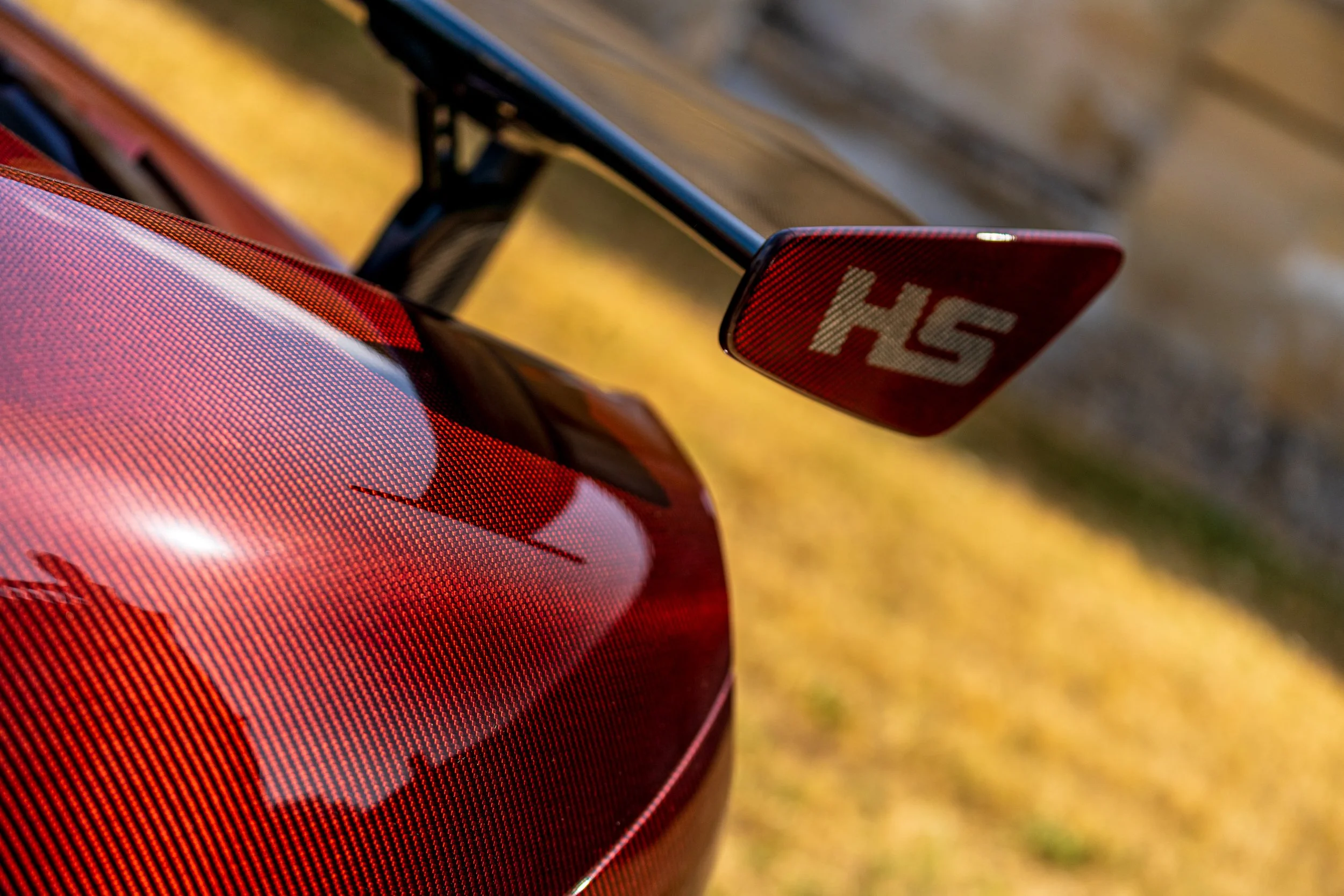 Close-up of red carbon mclaren's spoiler with 'HS' showing on the wing plate, parked on grass with blurred background.