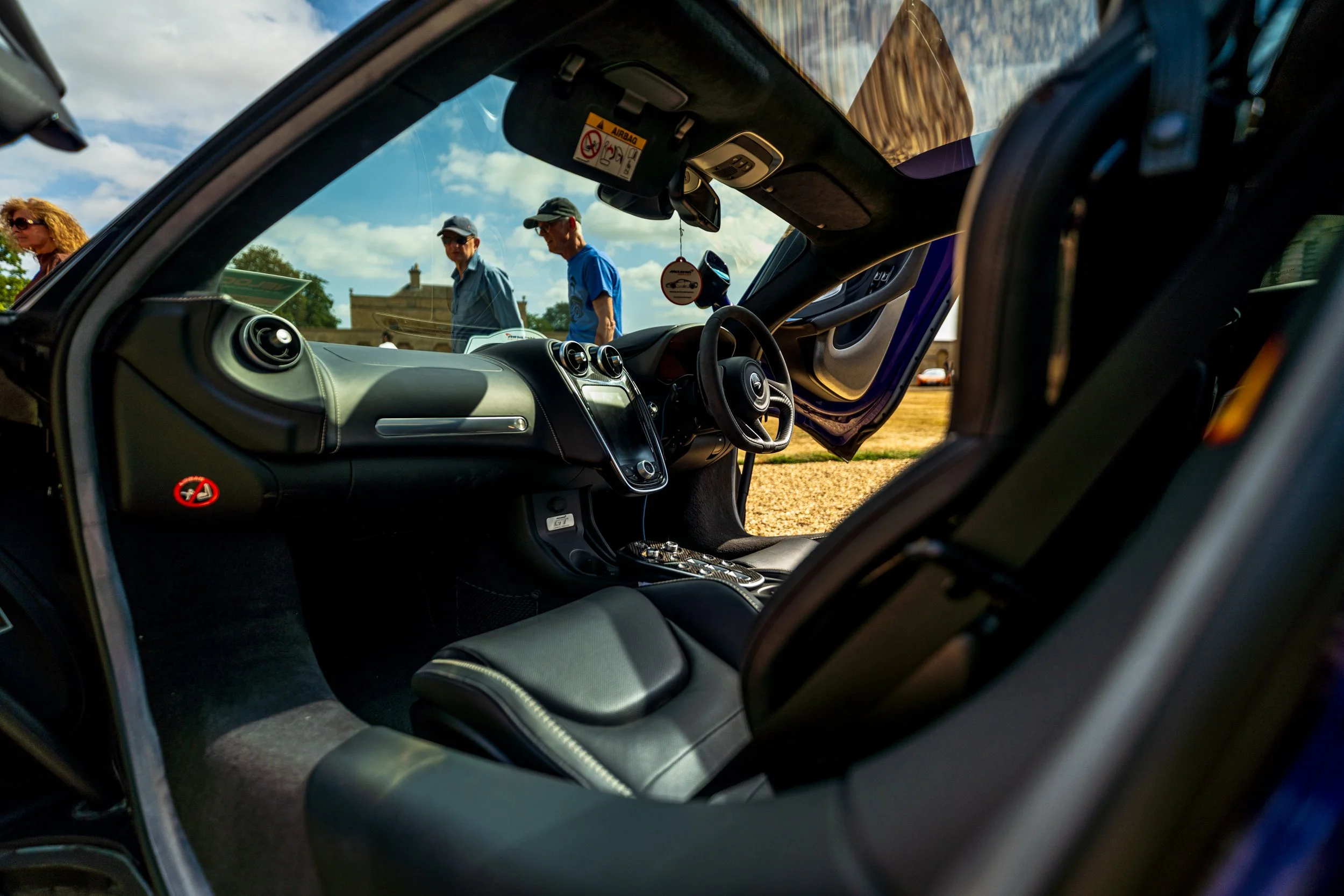 Interior of a car with the passenger door open, revealing the dashboard and steering wheel. Outside, two men and a woman are talking near the vehicle on a dirt or gravel surface, with a building and trees in the background and partly cloudy sky overh