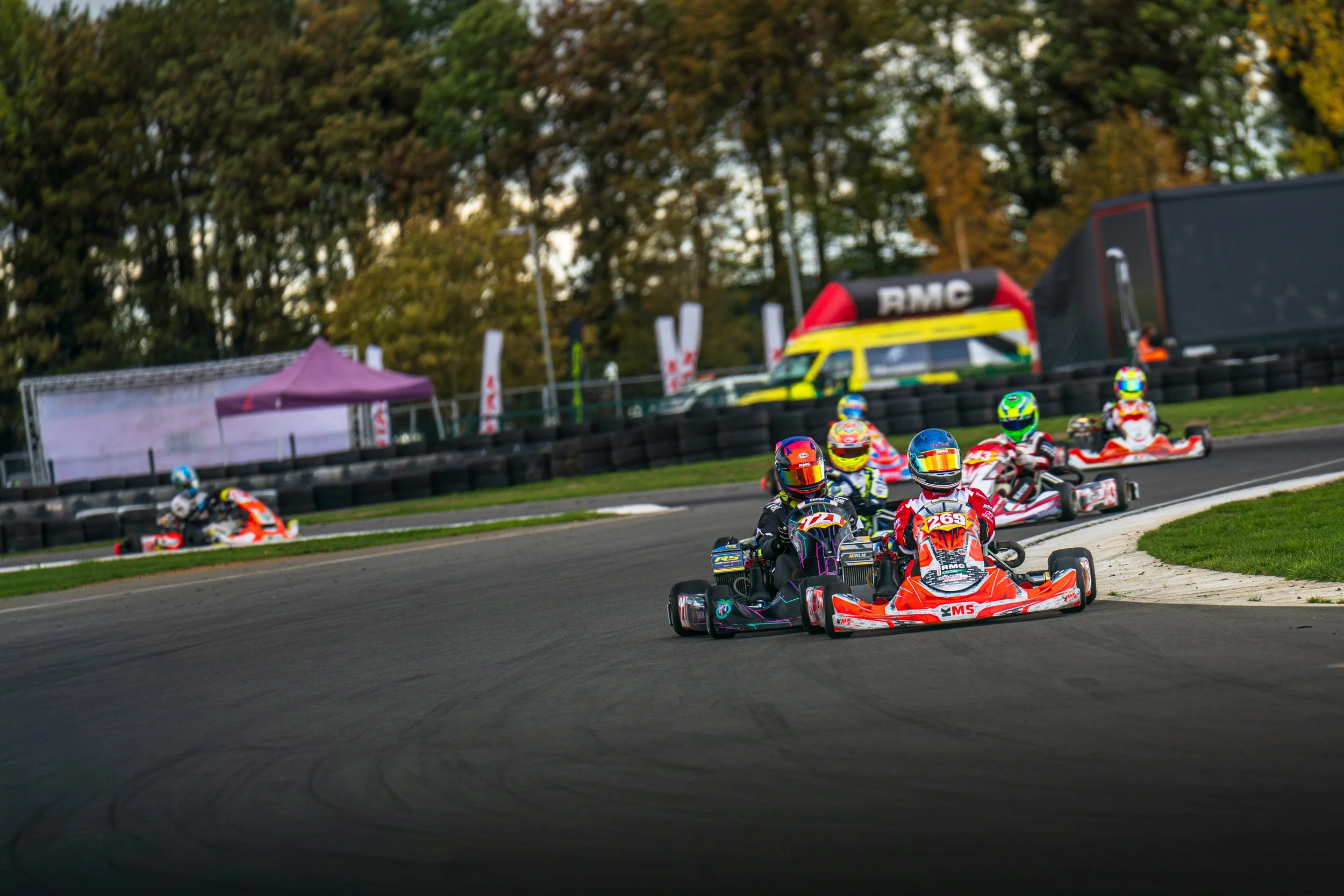 A field of colourful karts racing past trees at Silverstone.