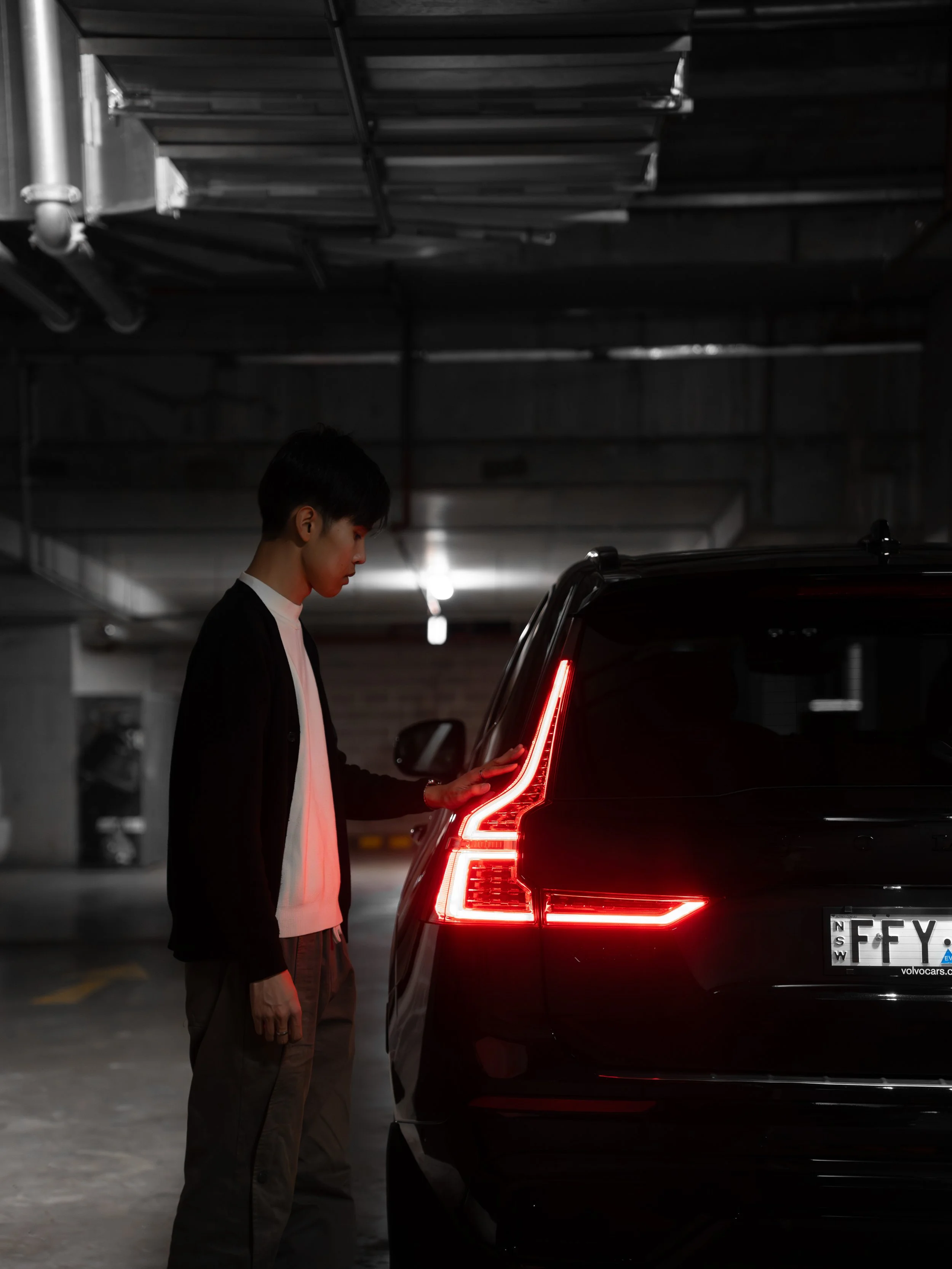 A young man in casual clothing looking at the taillight of a black car in an underground parking garage.