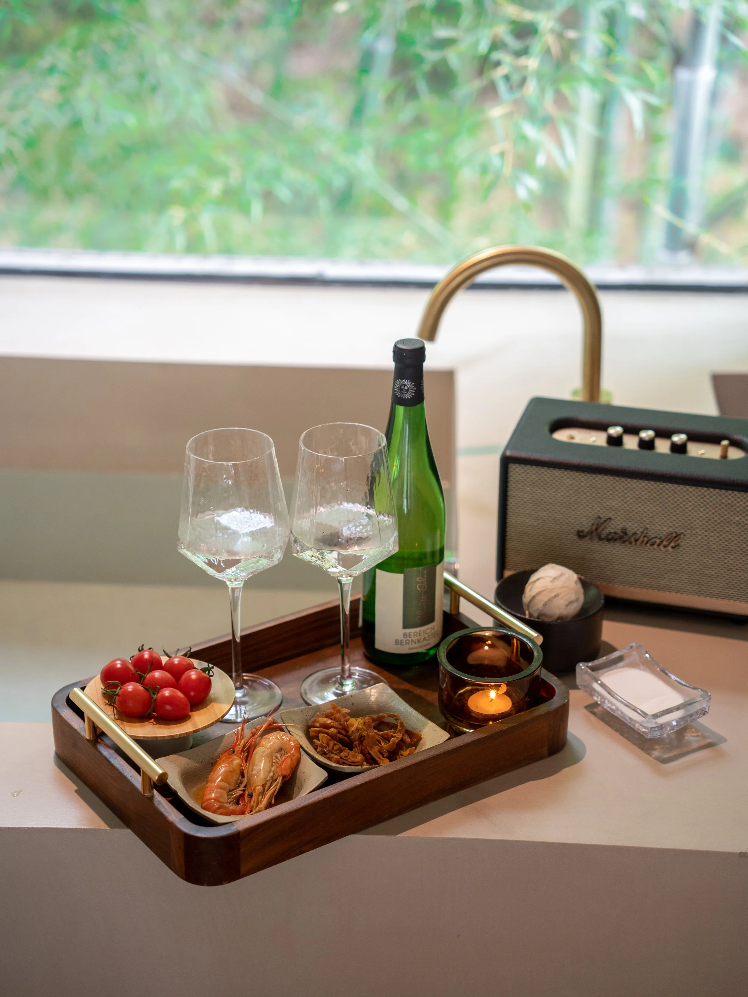 A wooden tray with seafood including shrimp, cherry tomatoes, and what appears to be fried or grilled fish, along with a bottle of sparkling wine, two wine glasses, a small candle holder, a Marshall speaker, a glass of whiskey, and various small dish