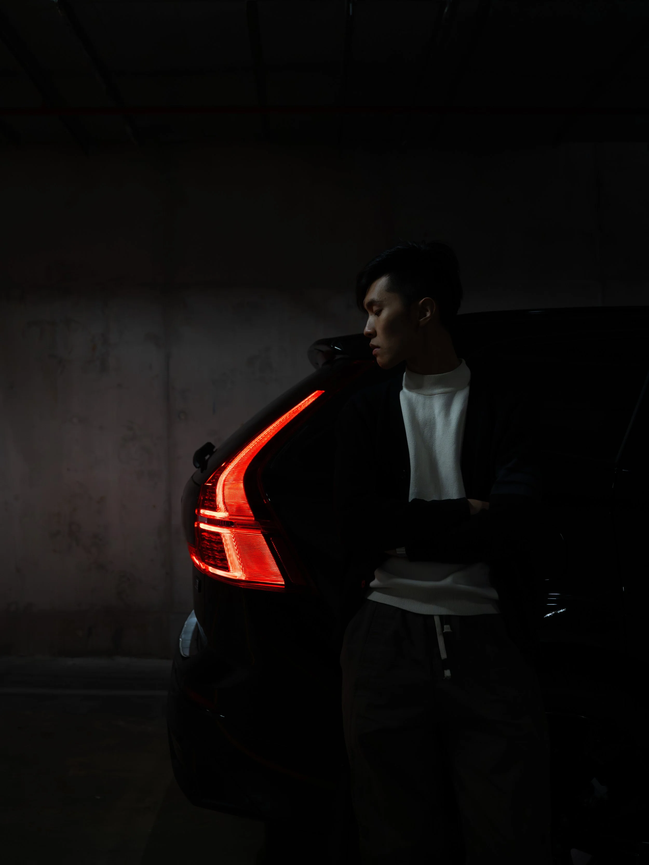 A young man with black hair standing next to a black car with illuminated tail lights in a dark, indoor parking garage.