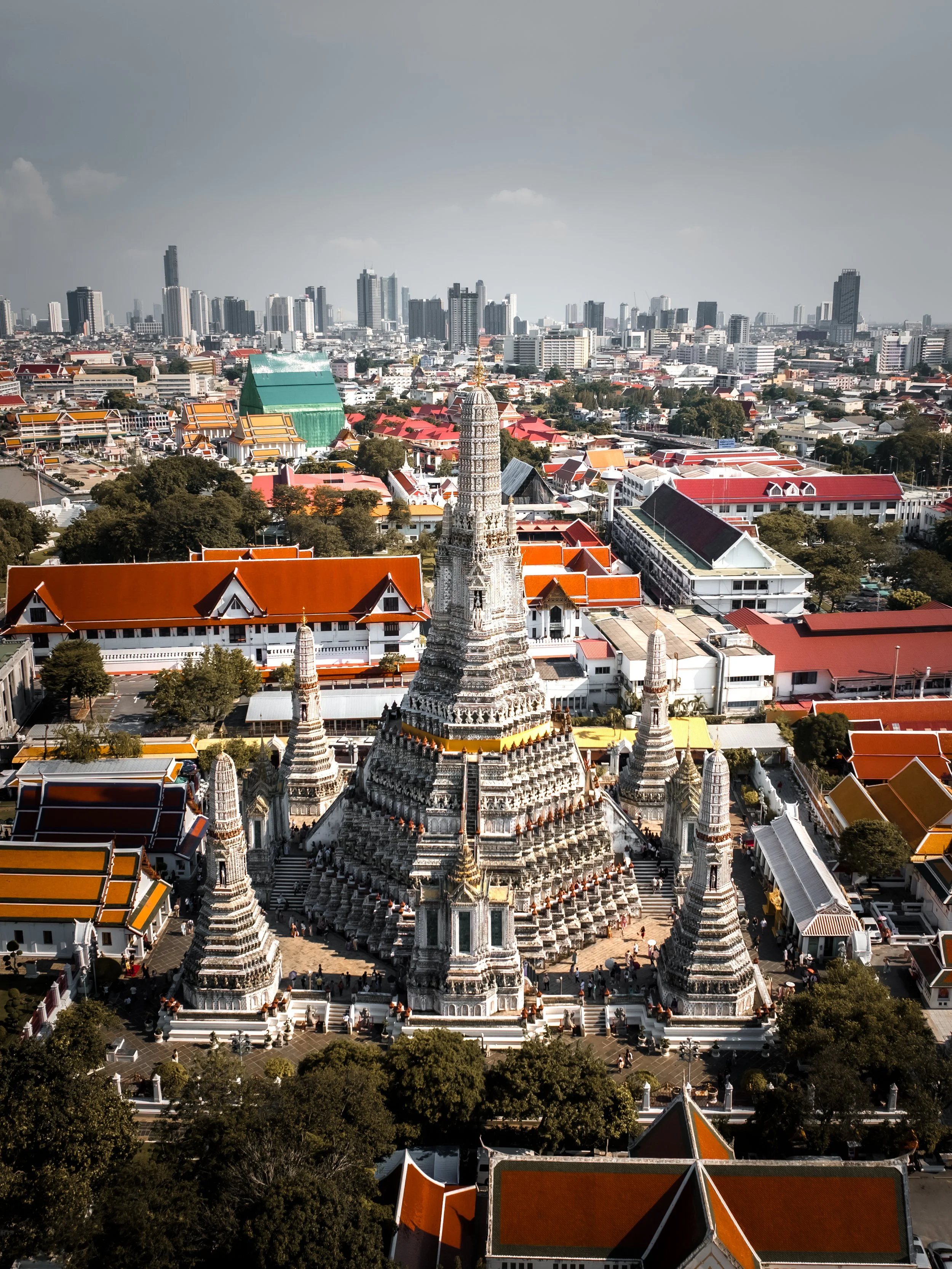 Aerial view of a cityscape with a historic temple in the foreground, featuring a central ornate white pagoda surrounded by smaller structures, with modern high-rise buildings in the background and trees throughout the area.