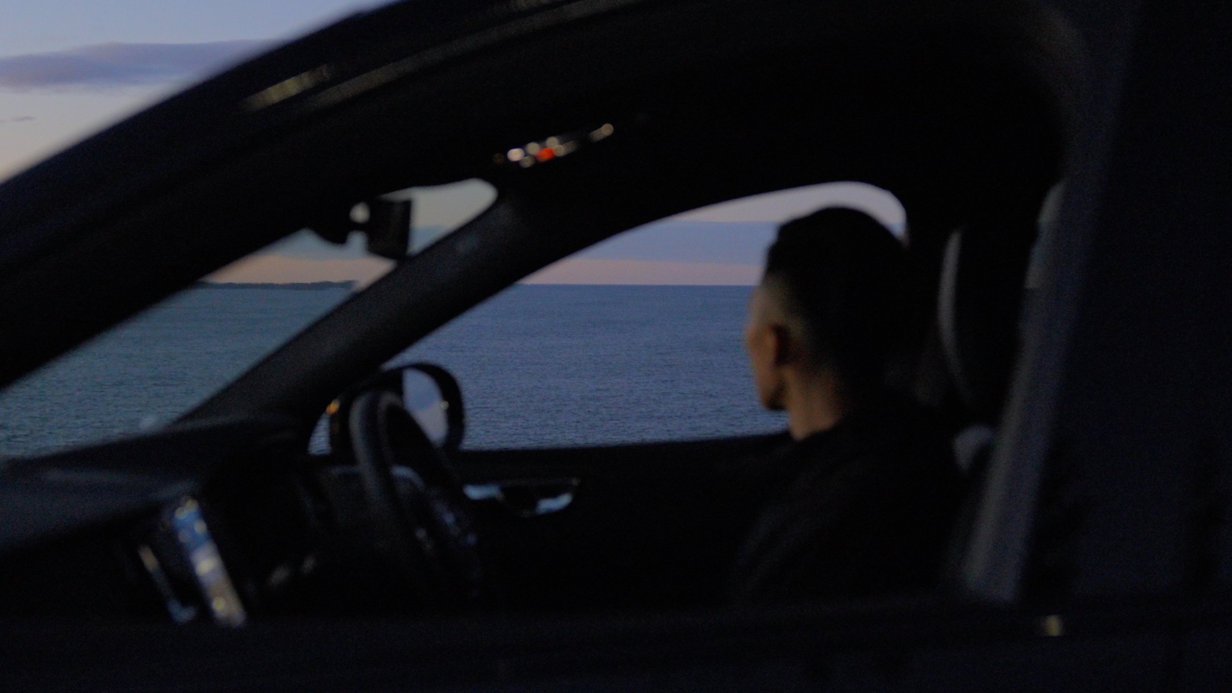 Inside a car, a person with dark hair is driving, visible from the side, with a view of the ocean and sky through the windshield.