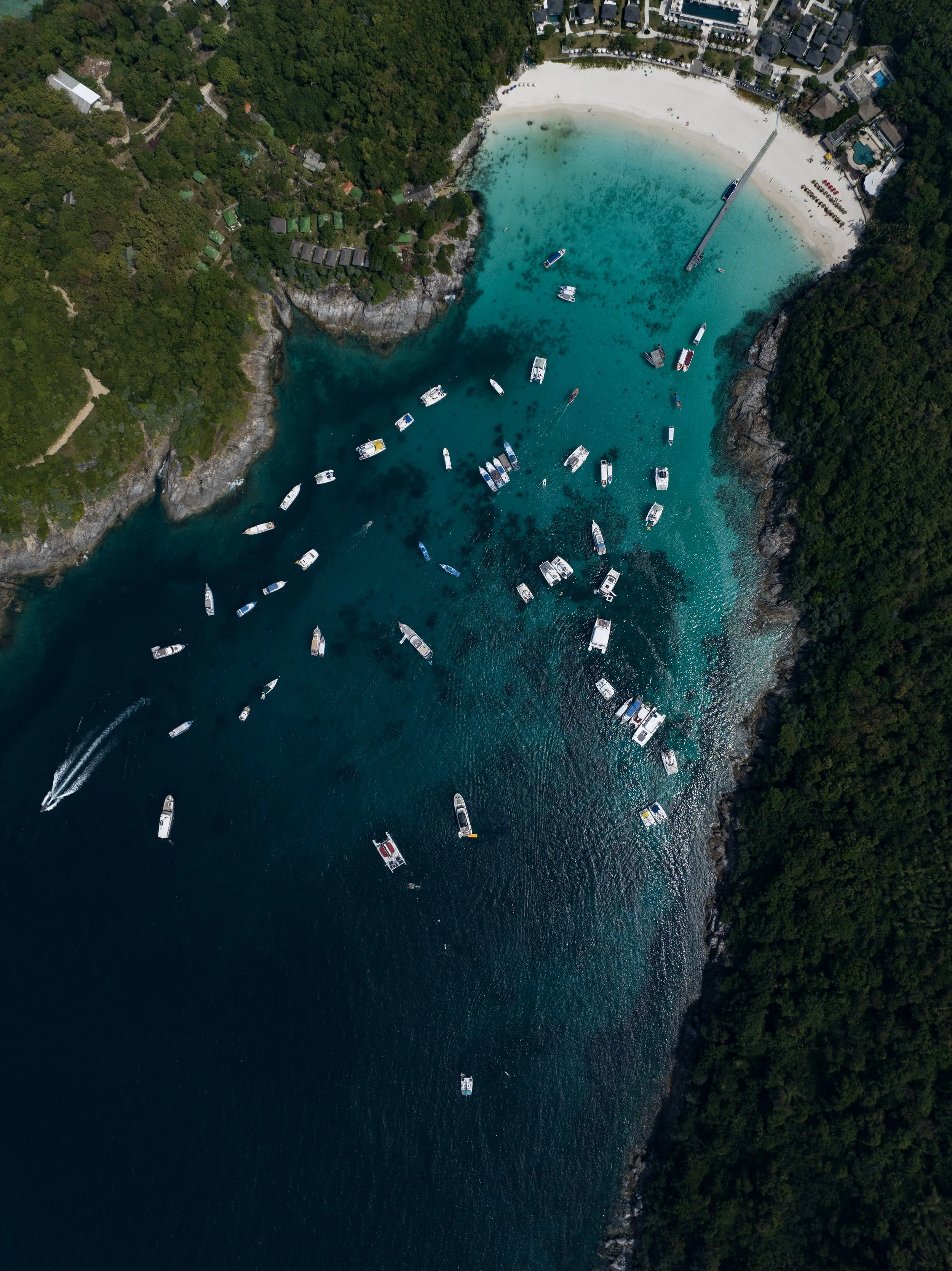 Aerial view of a coastal area featuring a sandy beach, turquoise water with boats, a pier, dense green forest, and resort buildings.