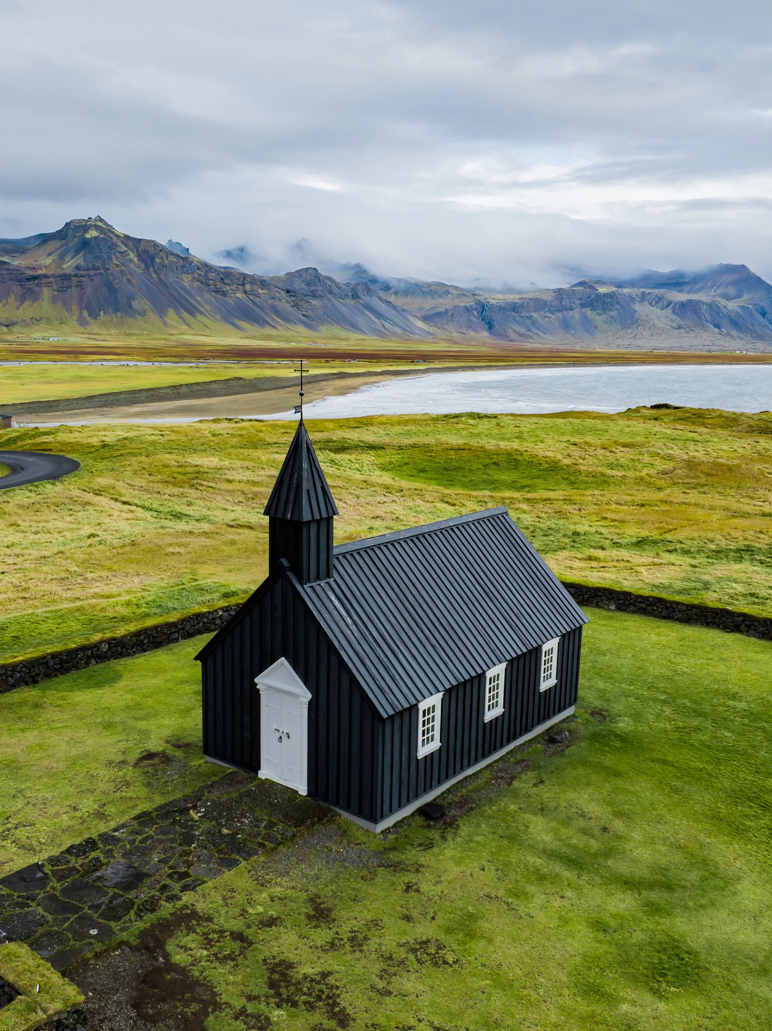 A black wooden church with a black roof and white door, set on a grassy landscape with mountains and a body of water in the background, under a cloudy sky.