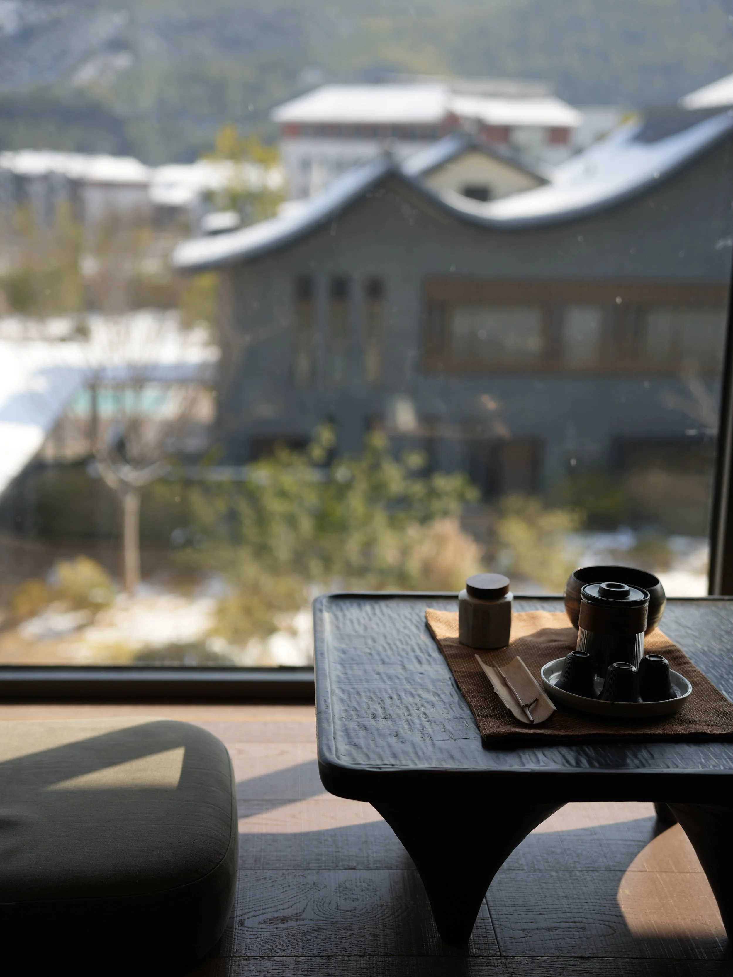 A table set for a meal with chopsticks and cups, positioned in front of a large window with a view of snow-covered roofs and trees outside.