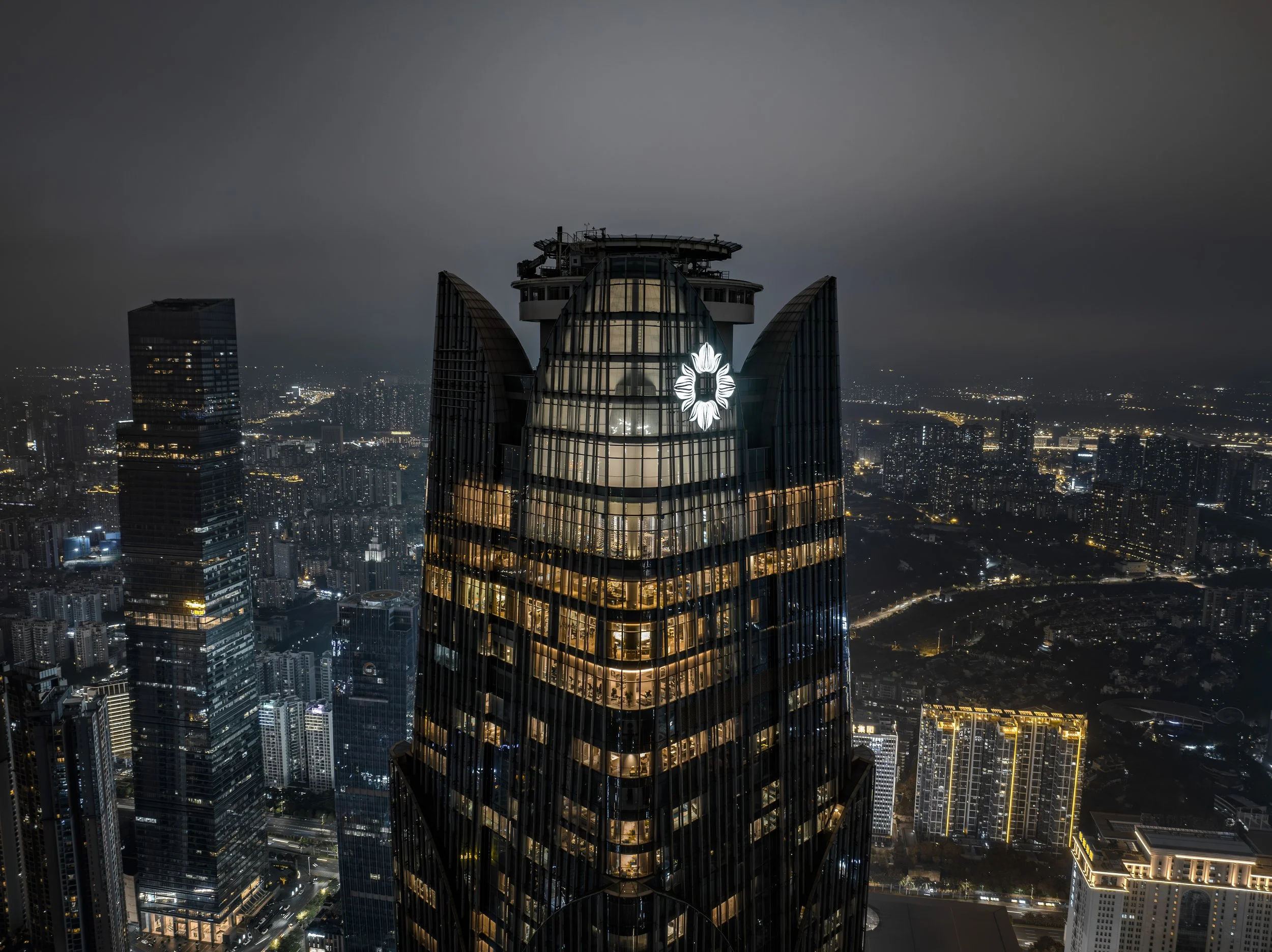 Nighttime cityscape with tall skyscraper in the foreground, featuring illuminated windows and a logo near the top. City lights and buildings extend into the distance under a cloudy sky.