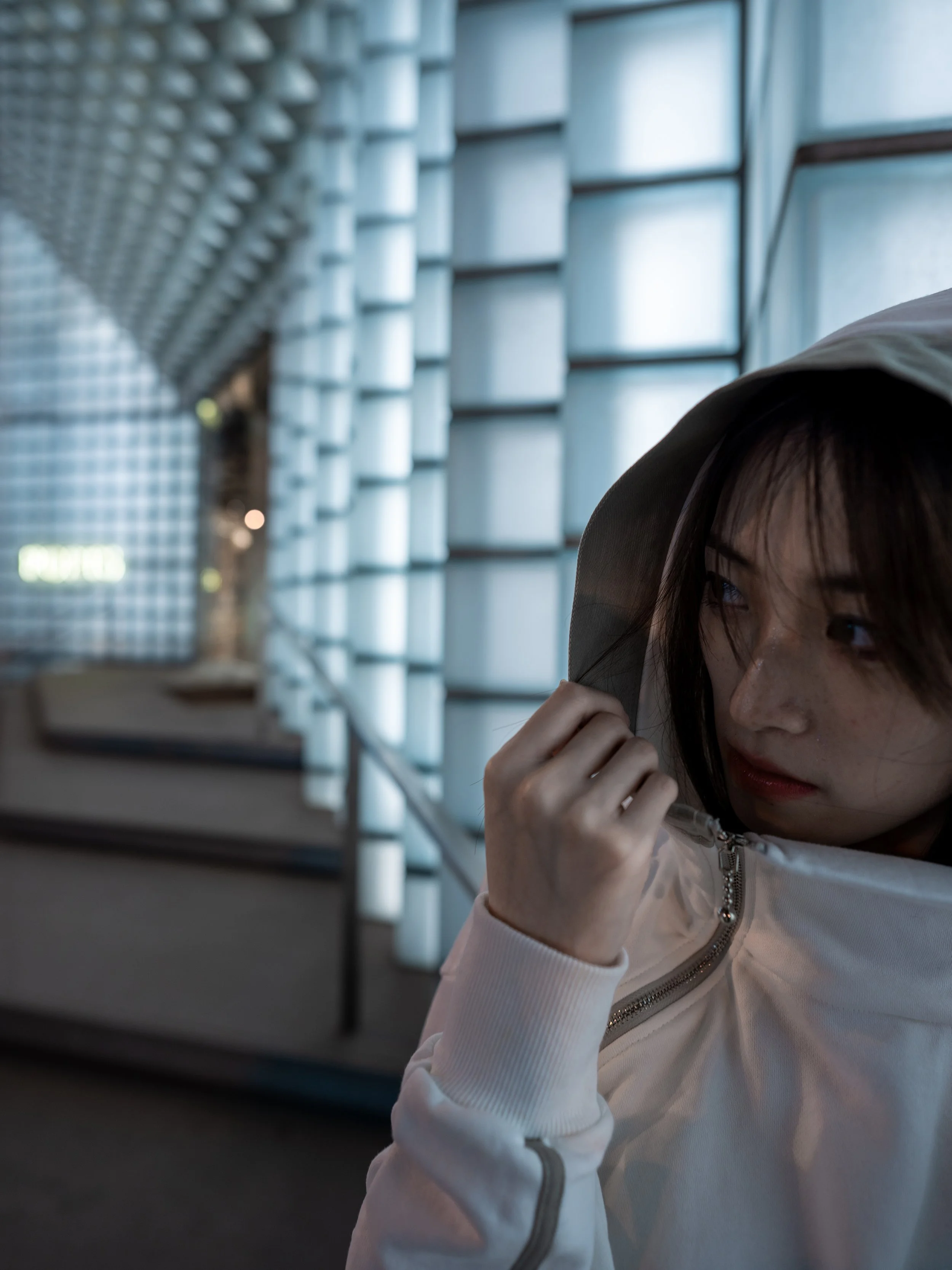 Close-up of a young girl with dark hair and freckles, wearing a white hoodie, holding a transparent umbrella, standing indoors near a large window with a grid design.